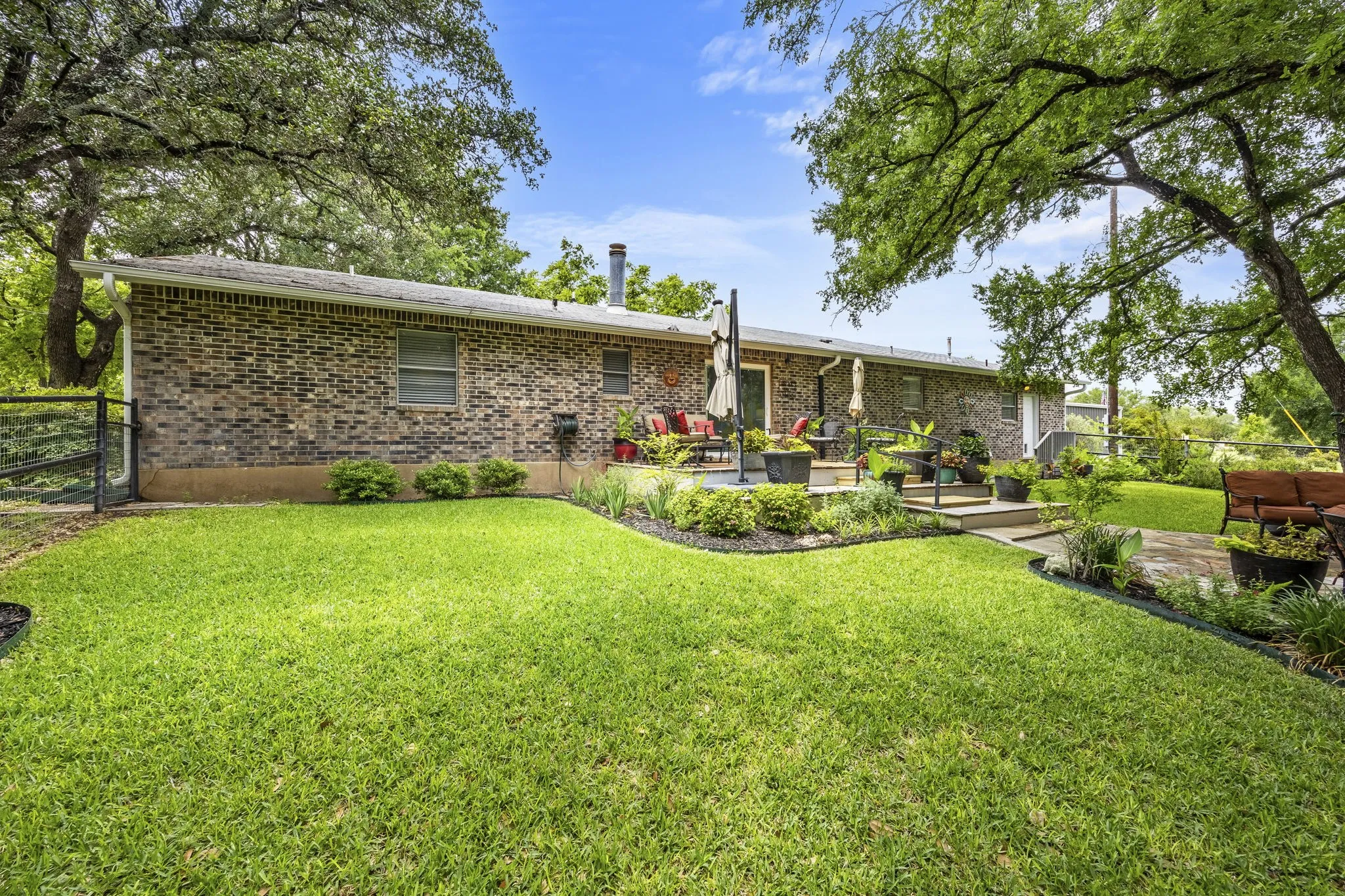 Expansive fenced backyard featuring a brick exterior, a multi-level patio with stone steps, mature trees, and a manicured lawn with defined garden beds