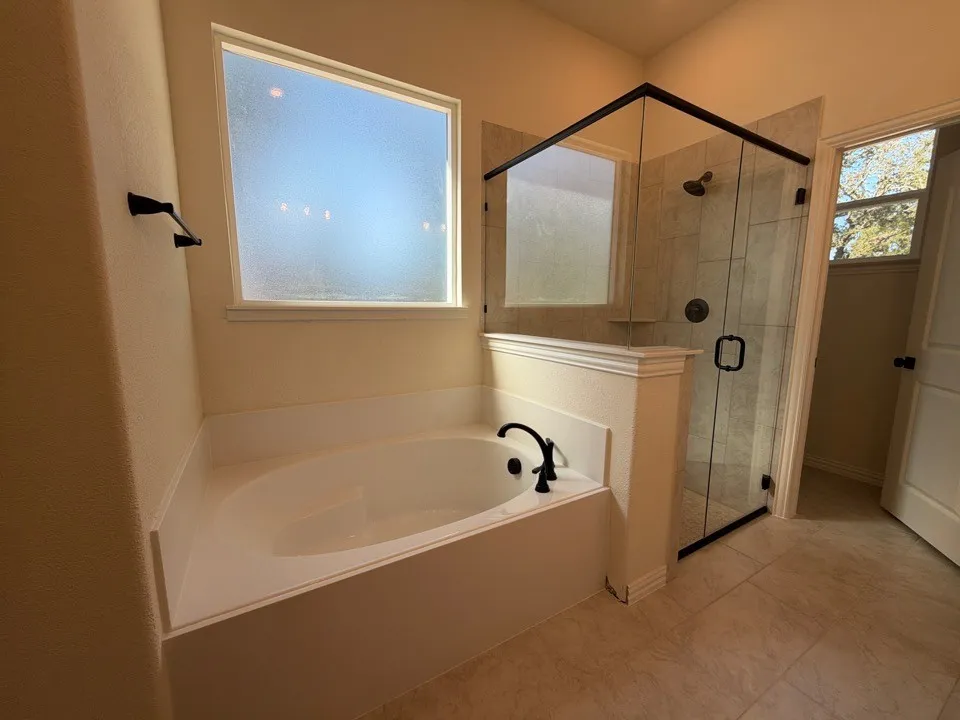 Soaking tub with black fixtures, tiled shower with glass enclosure, large privacy window, and neutral tile flooring
