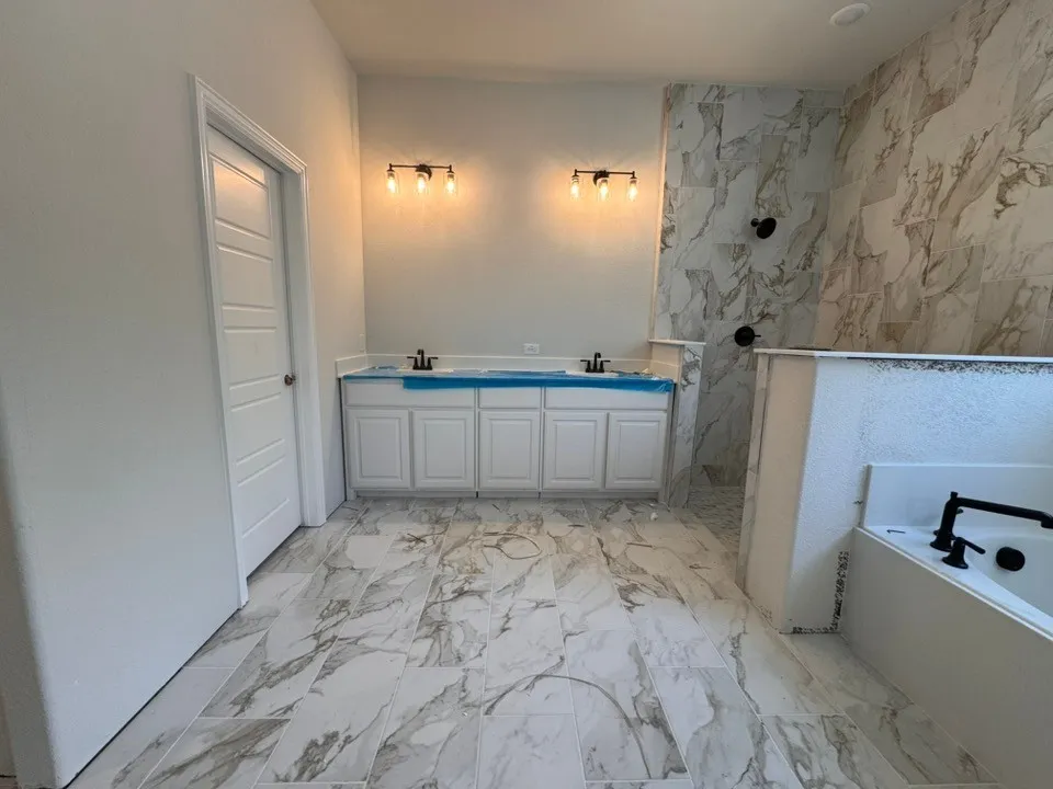 Dual vanity with white cabinetry, tile flooring, wall-mounted light fixtures, and a soaking tub