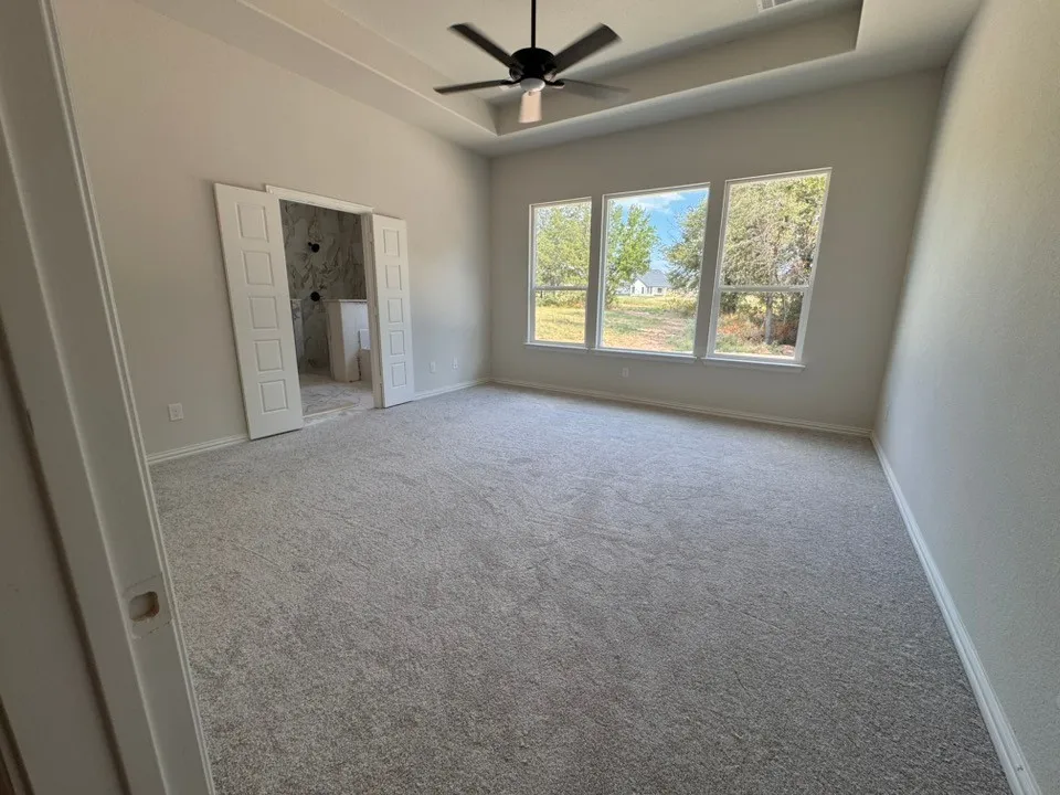 Spacious room featuring light-toned carpeting, a tray ceiling with a ceiling fan, and three large windows