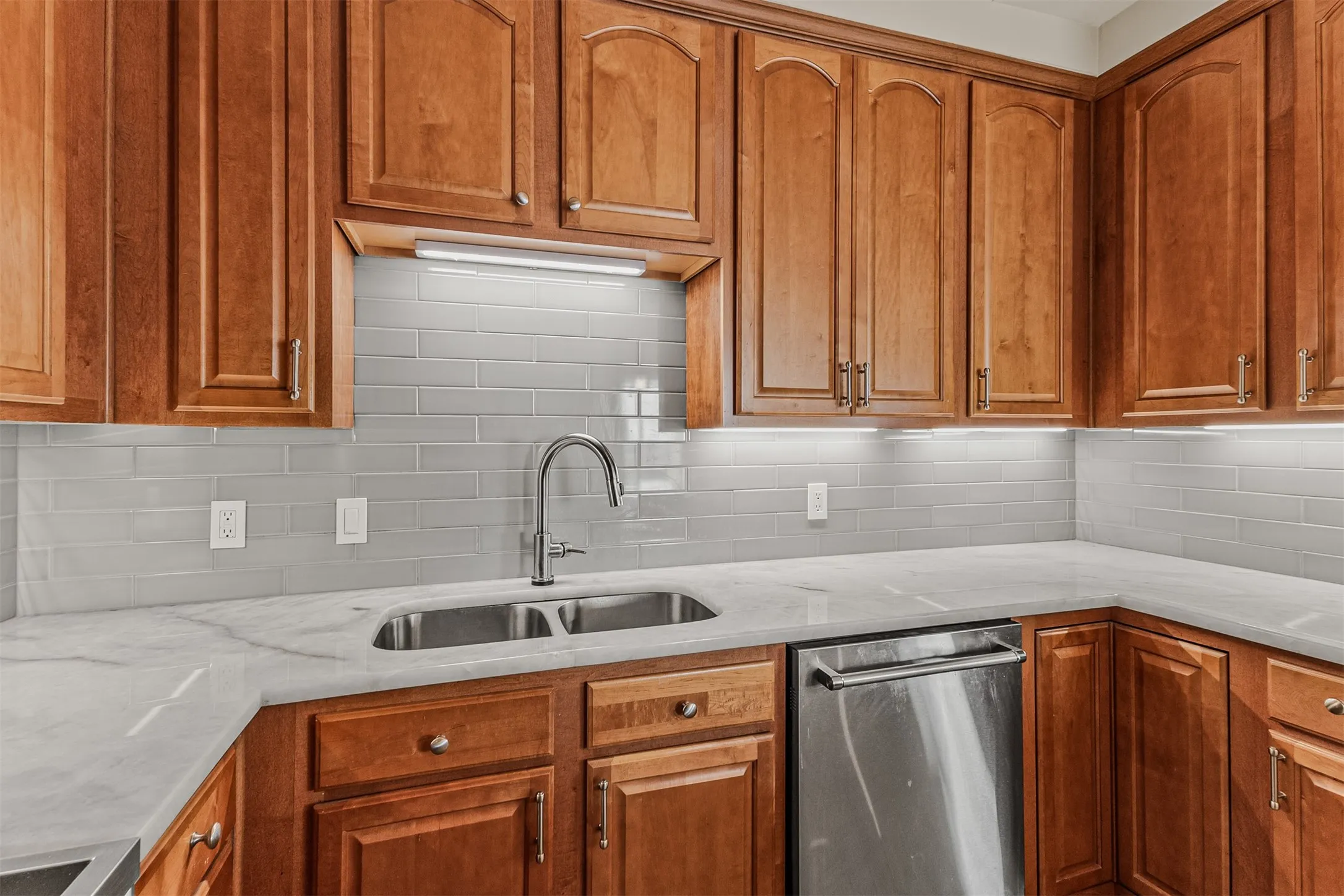 Kitchen featuring wood-finish cabinetry, light-toned countertops, a subway tile backsplash, a double basin sink with a gooseneck faucet, and a stainless steel dishwasher