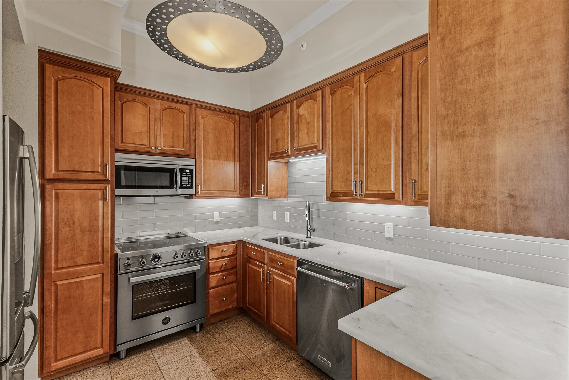 Kitchen featuring wood cabinetry, stainless steel appliances, white subway tile backsplash, light-toned countertops, and a decorative ceiling light fixture