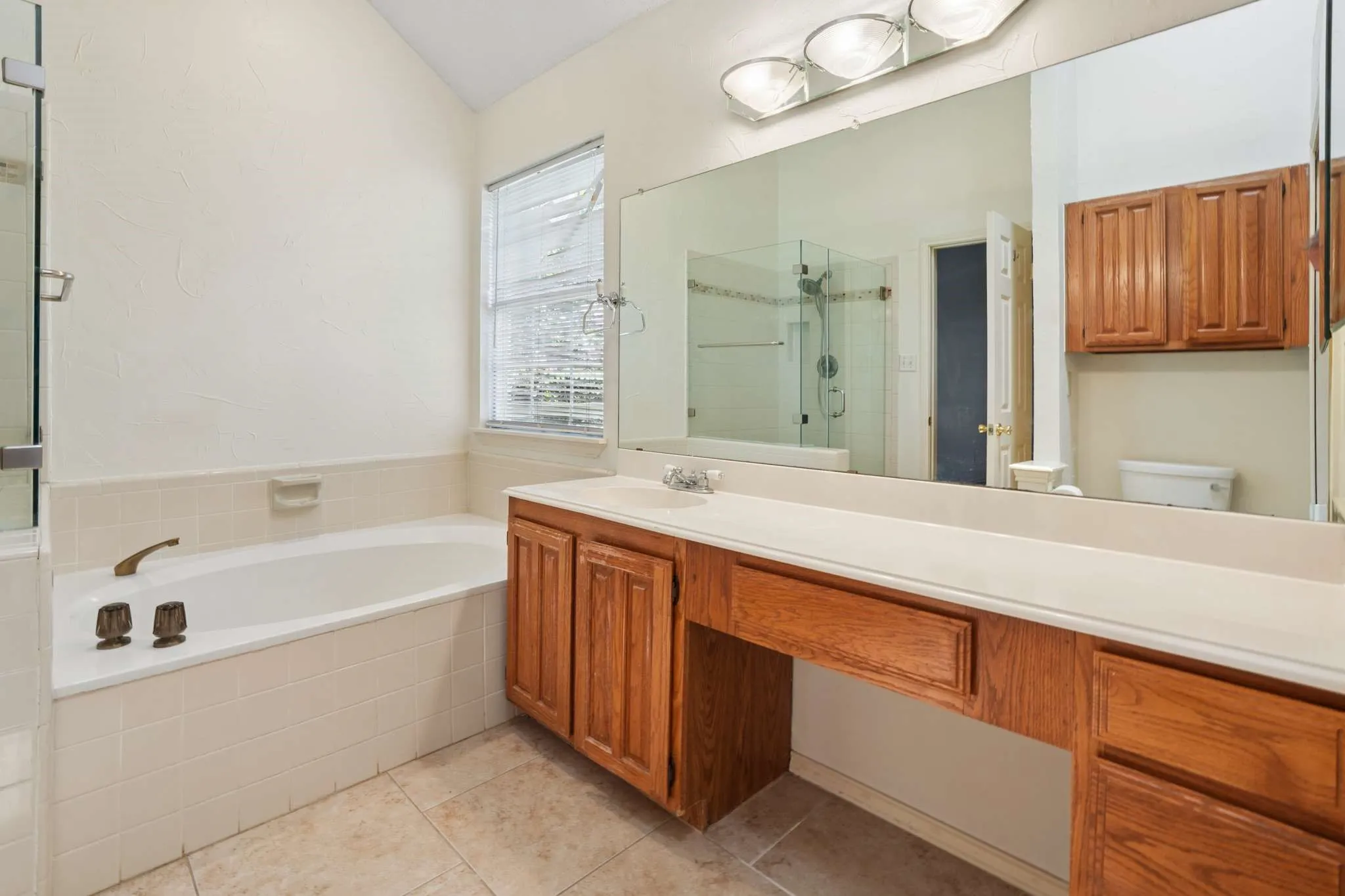 Bathroom featuring a built-in bathtub with tiled surround, a separate glass-enclosed shower, wood-finish vanity cabinetry with an integrated countertop, a full-width mirror, and tile flooring