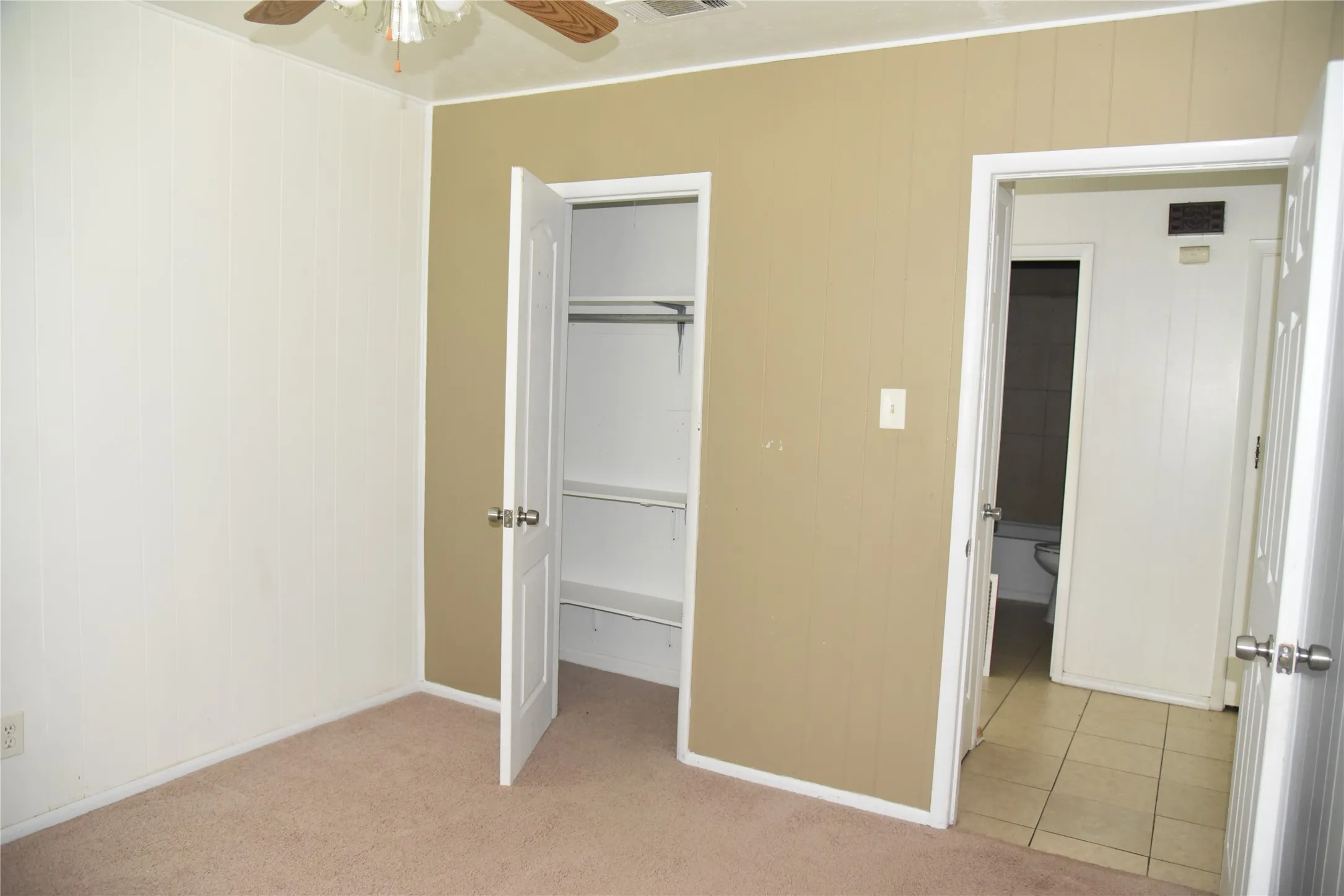 Carpeted room with vertical paneling on walls, featuring a ceiling fan, a built-in closet with shelving and hanging rod, and a white paneled door