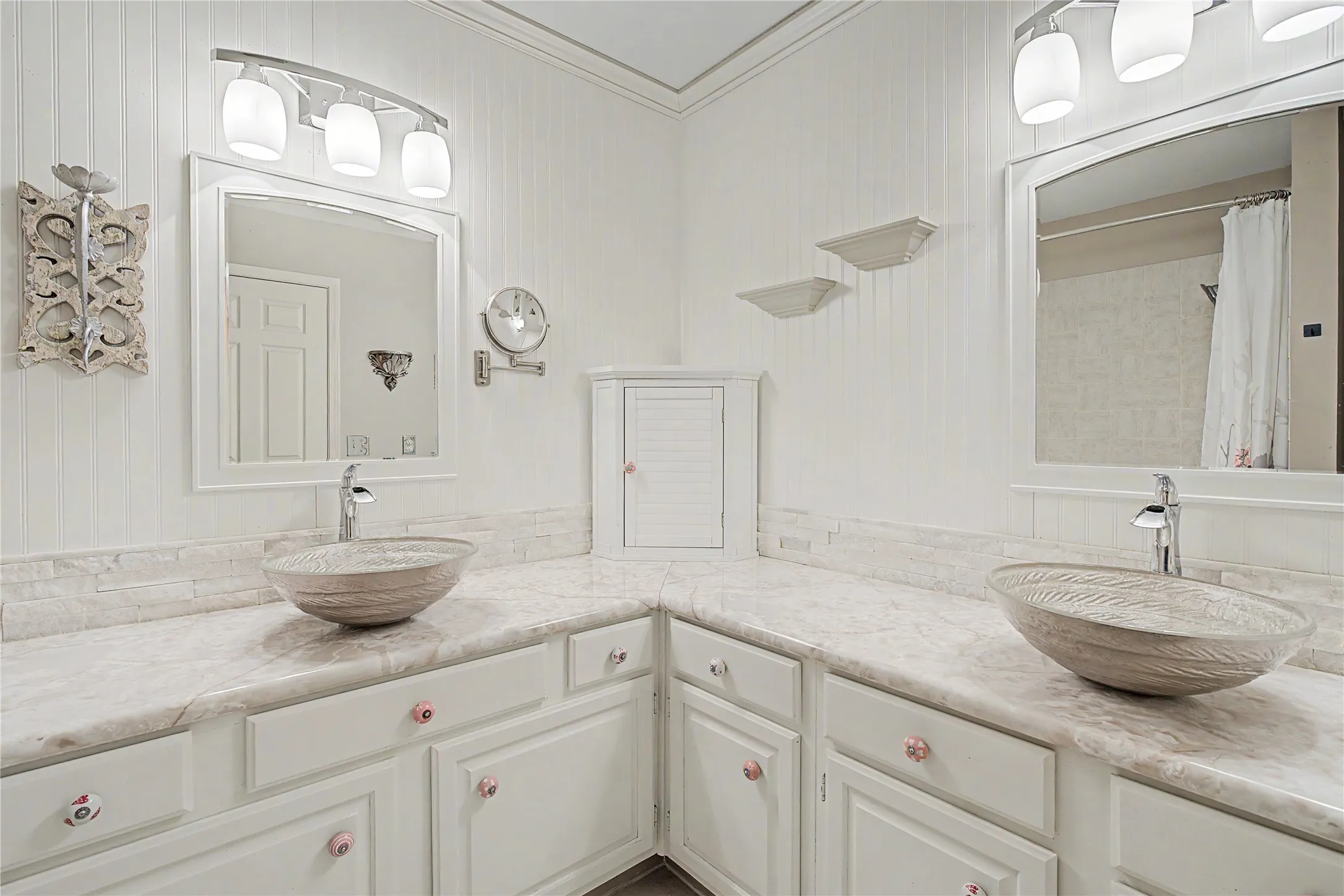 Dual vessel sinks with integrated chrome faucets, white cabinetry featuring decorative knobs, extensive countertop space with a matching backsplash, framed mirrors, and vertical paneled wall detailing