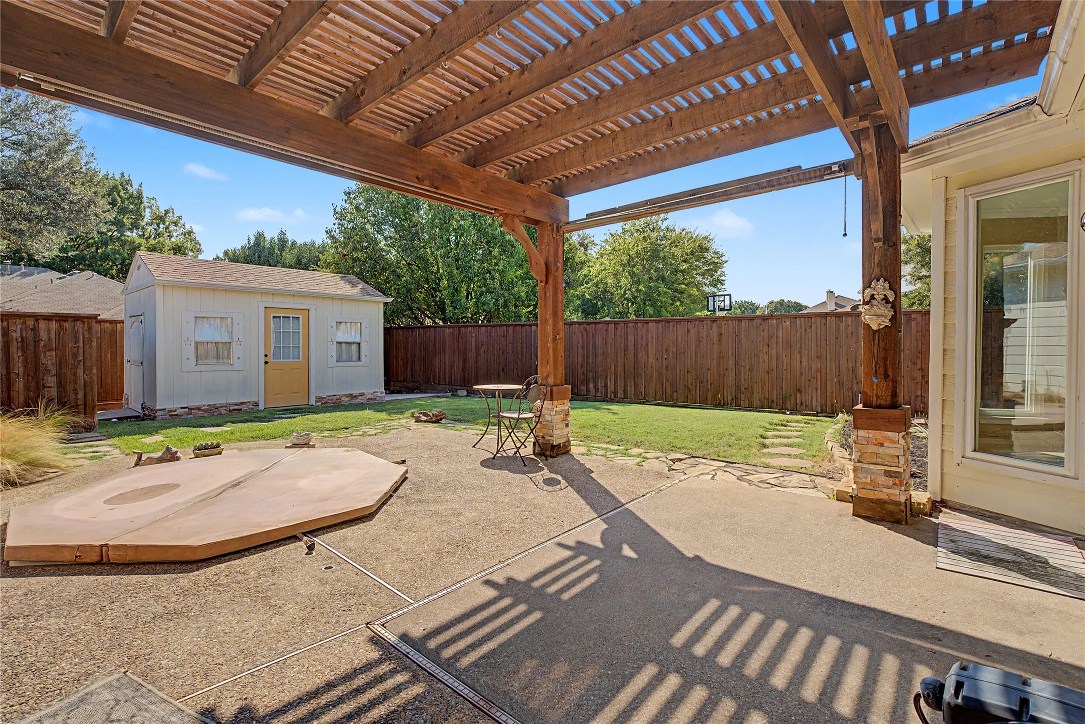Expansive patio featuring a wooden pergola with stone-clad pillars, concrete paving, a private wooden fence, and a storage shed