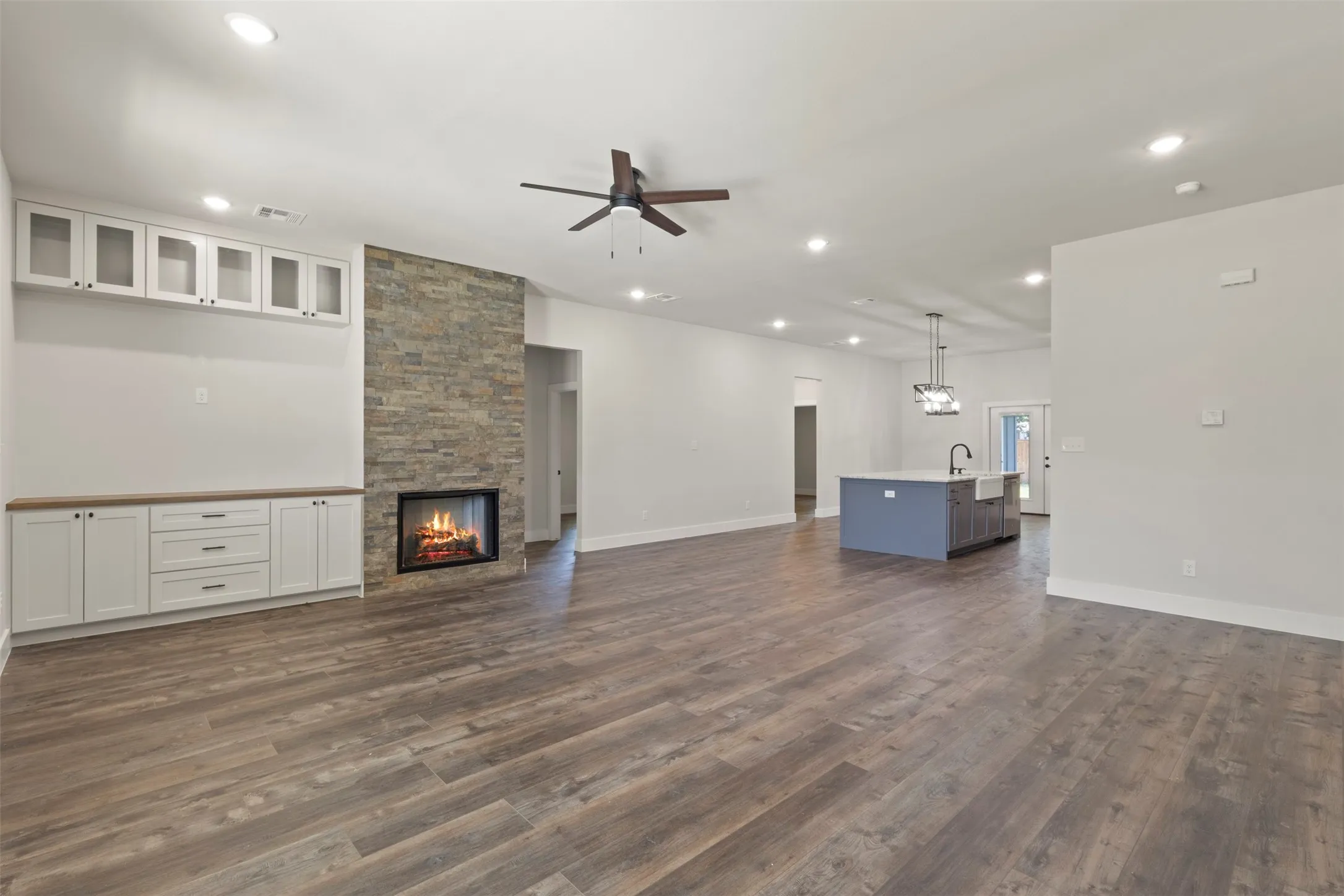 Open-concept living area with wood-finish flooring, a stacked stone fireplace, and custom built-in cabinetry featuring glass-front uppers and solid base storage