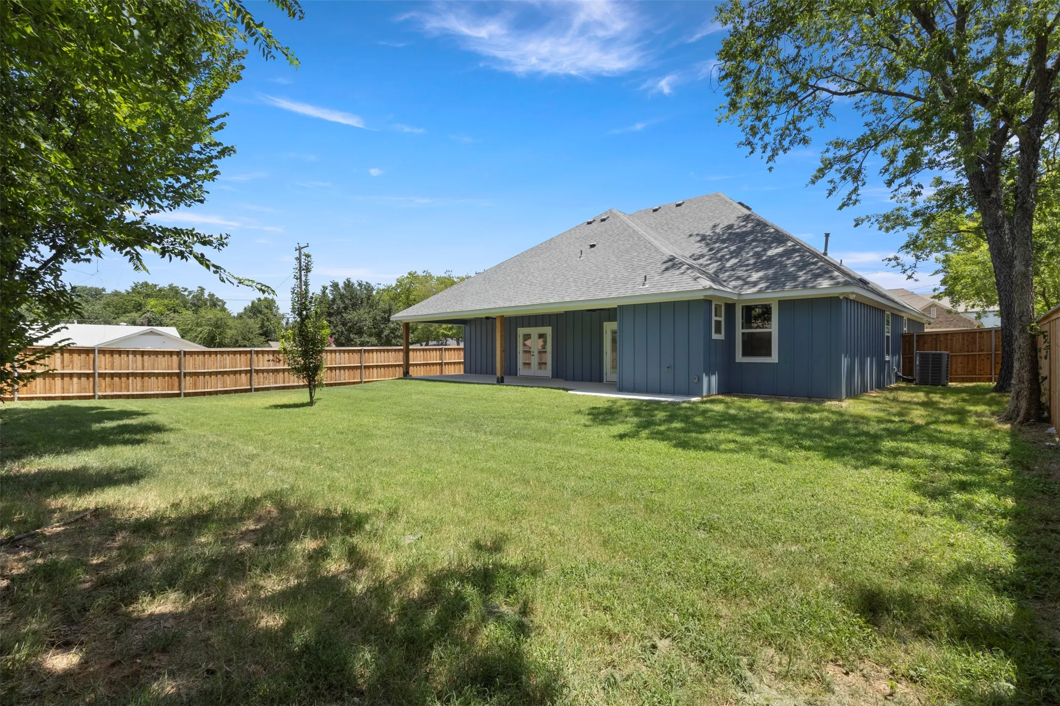 Expansive backyard featuring a manicured lawn, full privacy fencing, mature trees, a covered patio, and a light blue exterior