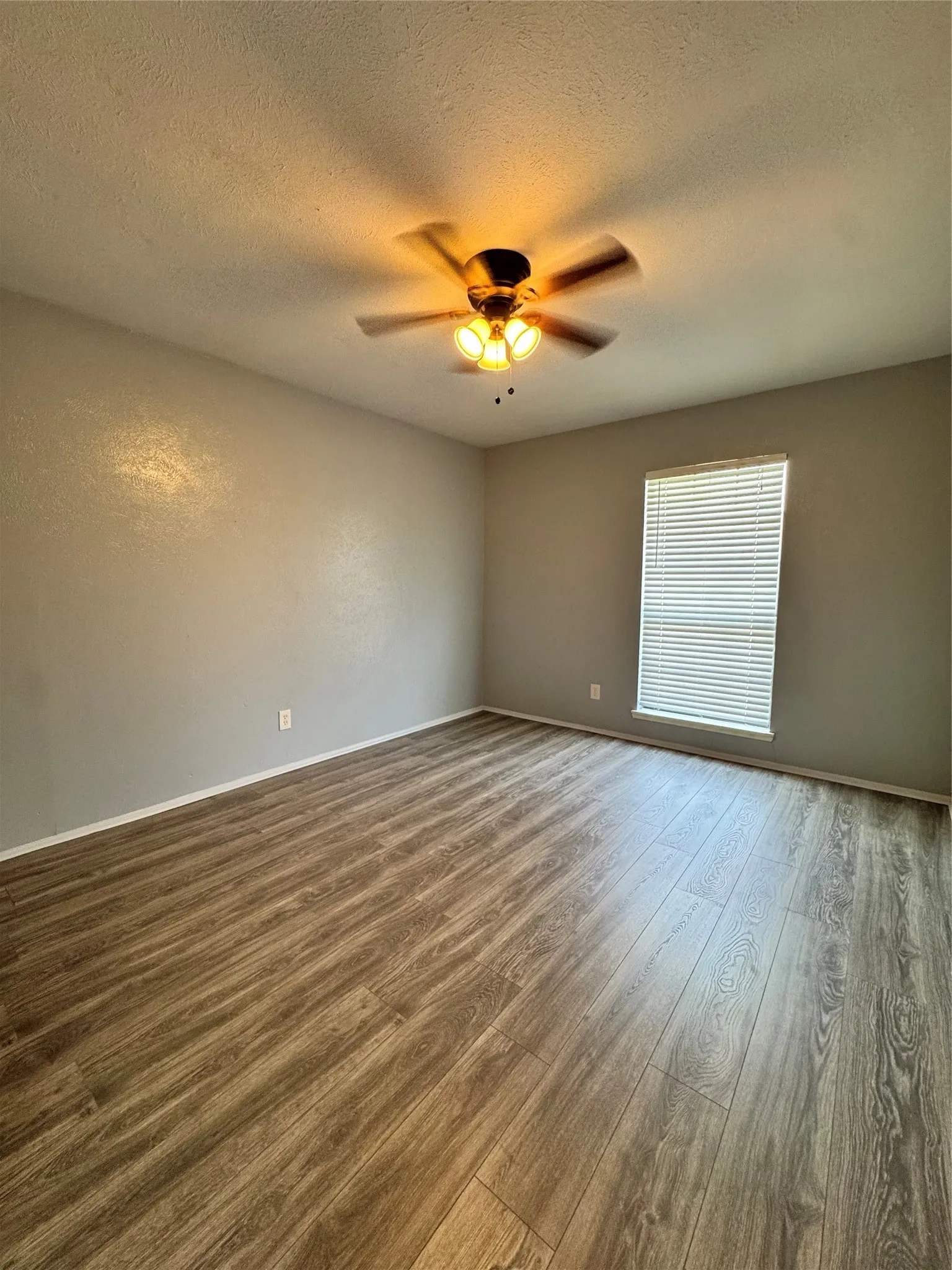 Spacious room with wood-finish flooring, a ceiling fan with integrated lighting, a single window with horizontal blinds, and neutral wall tones