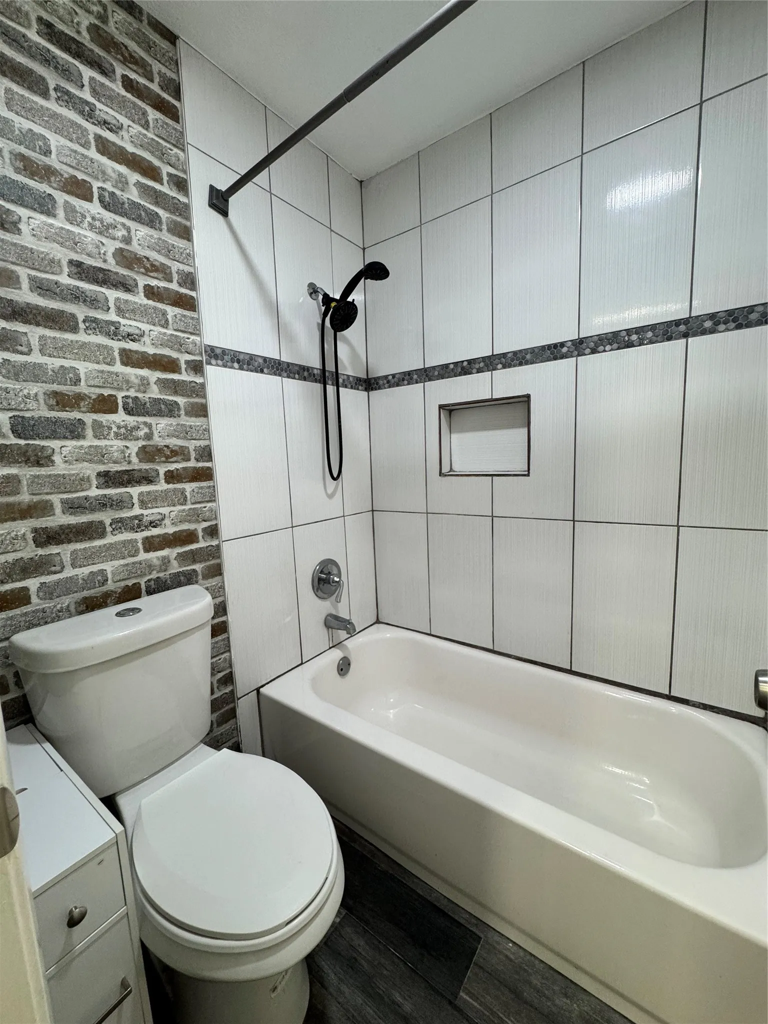 Bathroom featuring a white bathtub, white ceramic tile shower surround with a decorative mosaic band, integrated shower niche, and a contrasting brick-look accent wall