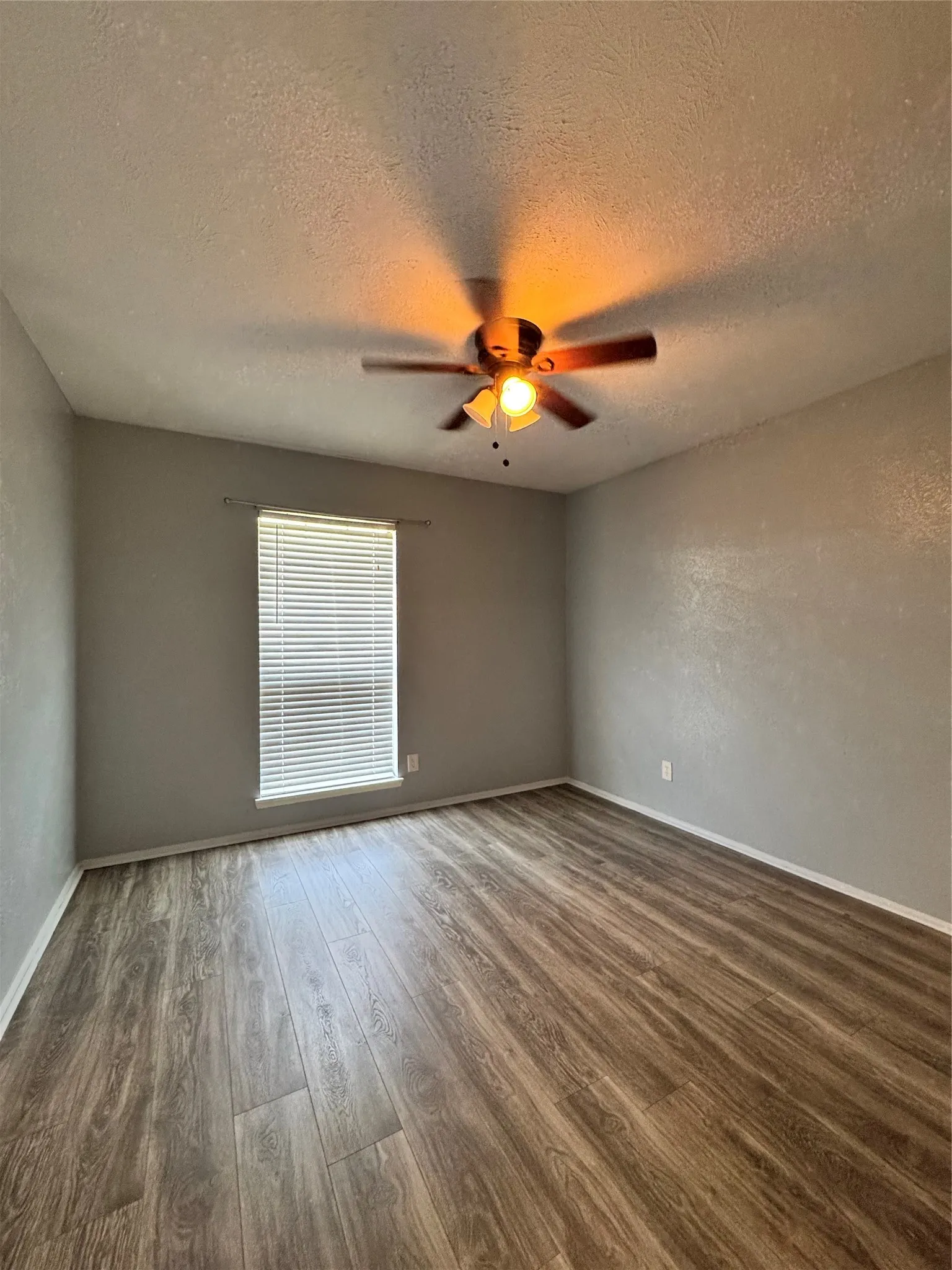 Spacious room featuring wood-finish flooring, light grey painted walls, a window with horizontal blinds, and a ceiling fan with integrated lighting