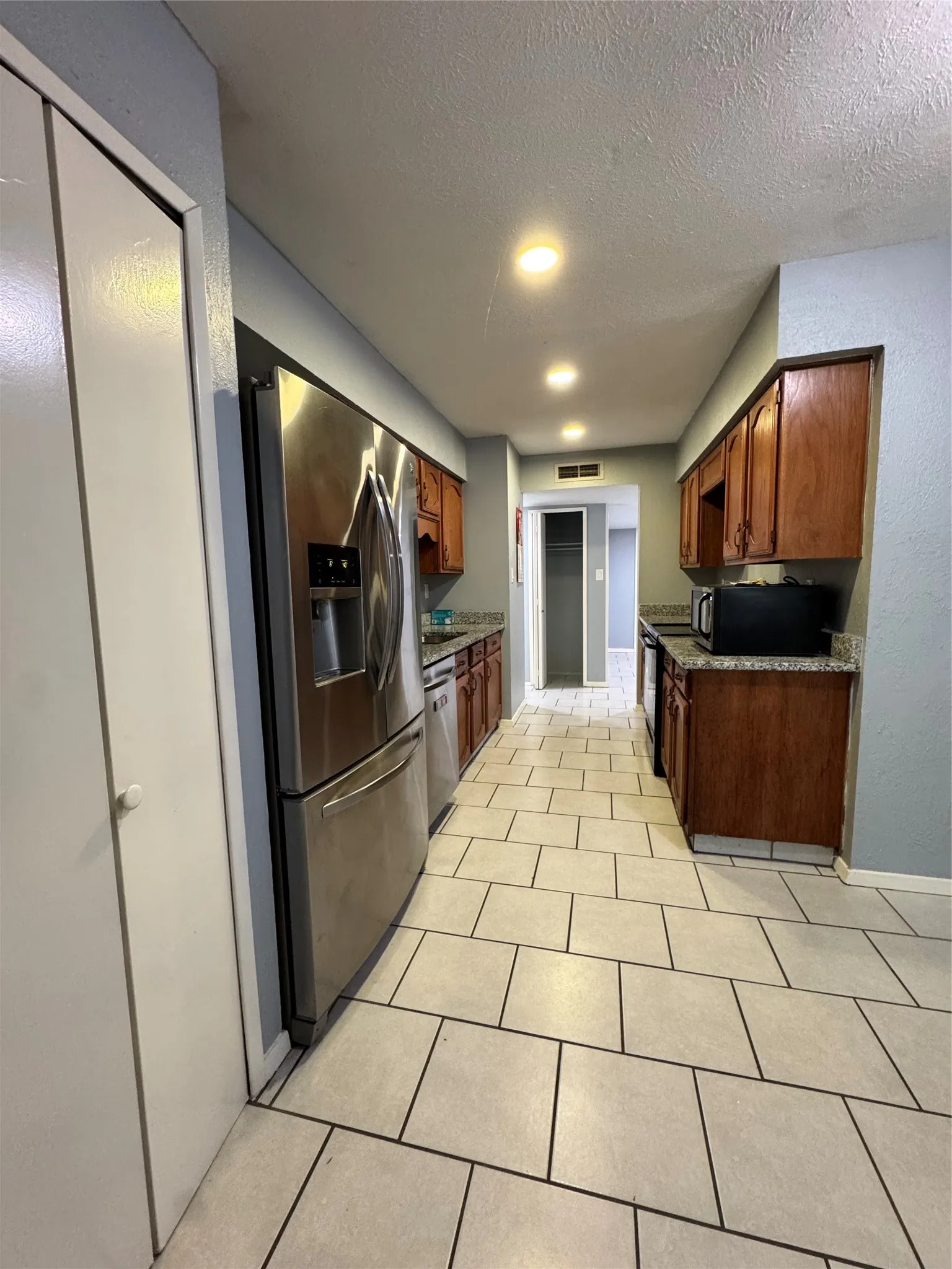 Galley kitchen featuring stainless steel appliances, recessed ceiling lighting, light-toned tile flooring, wood cabinetry, and speckled countertops