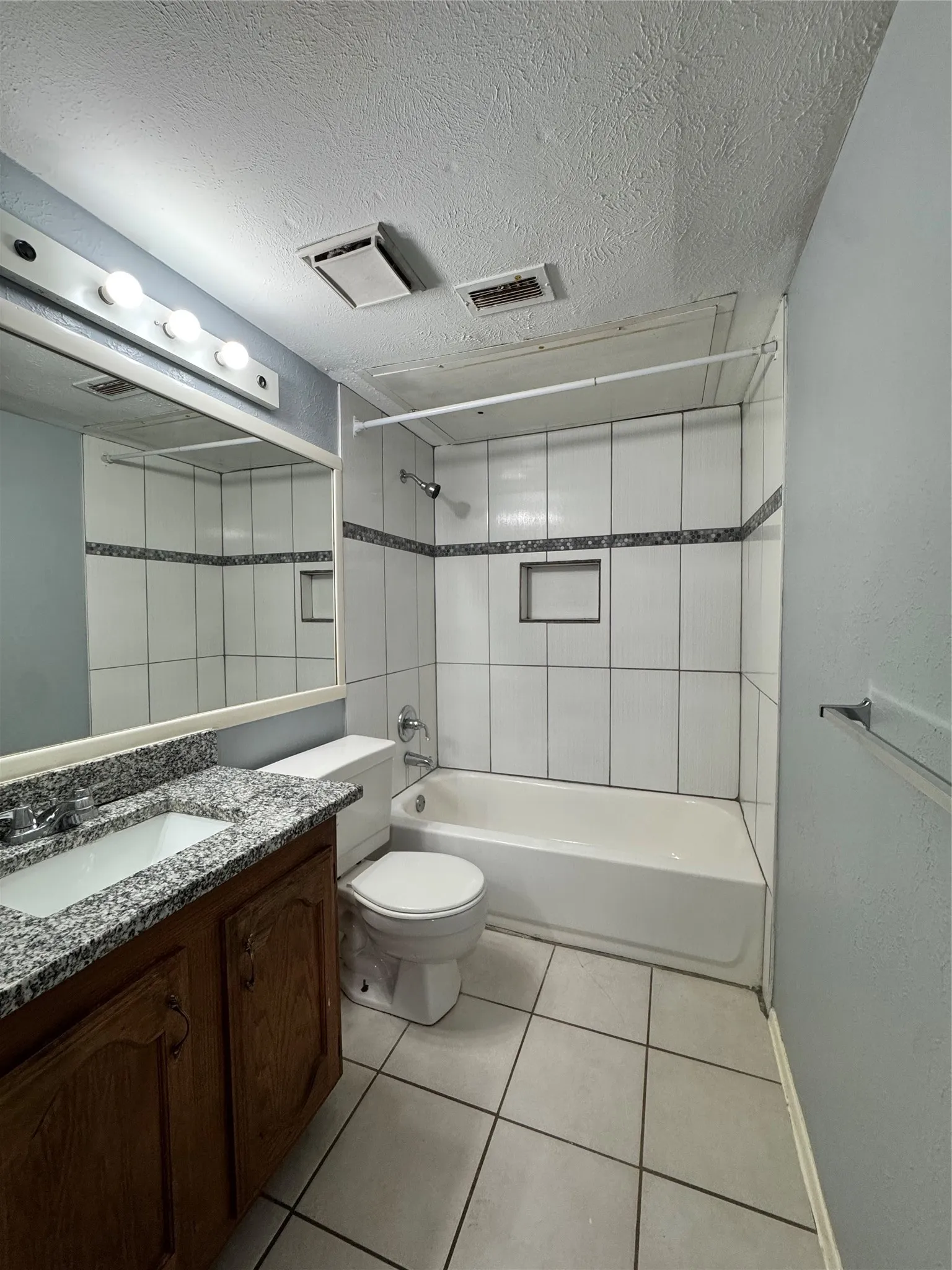 Bathroom featuring a tub/shower combination with white vertical tile and decorative accent band, a built-in shower niche, a vanity with a granite-patterned countertop and undermount sink, and light-toned floor tiles