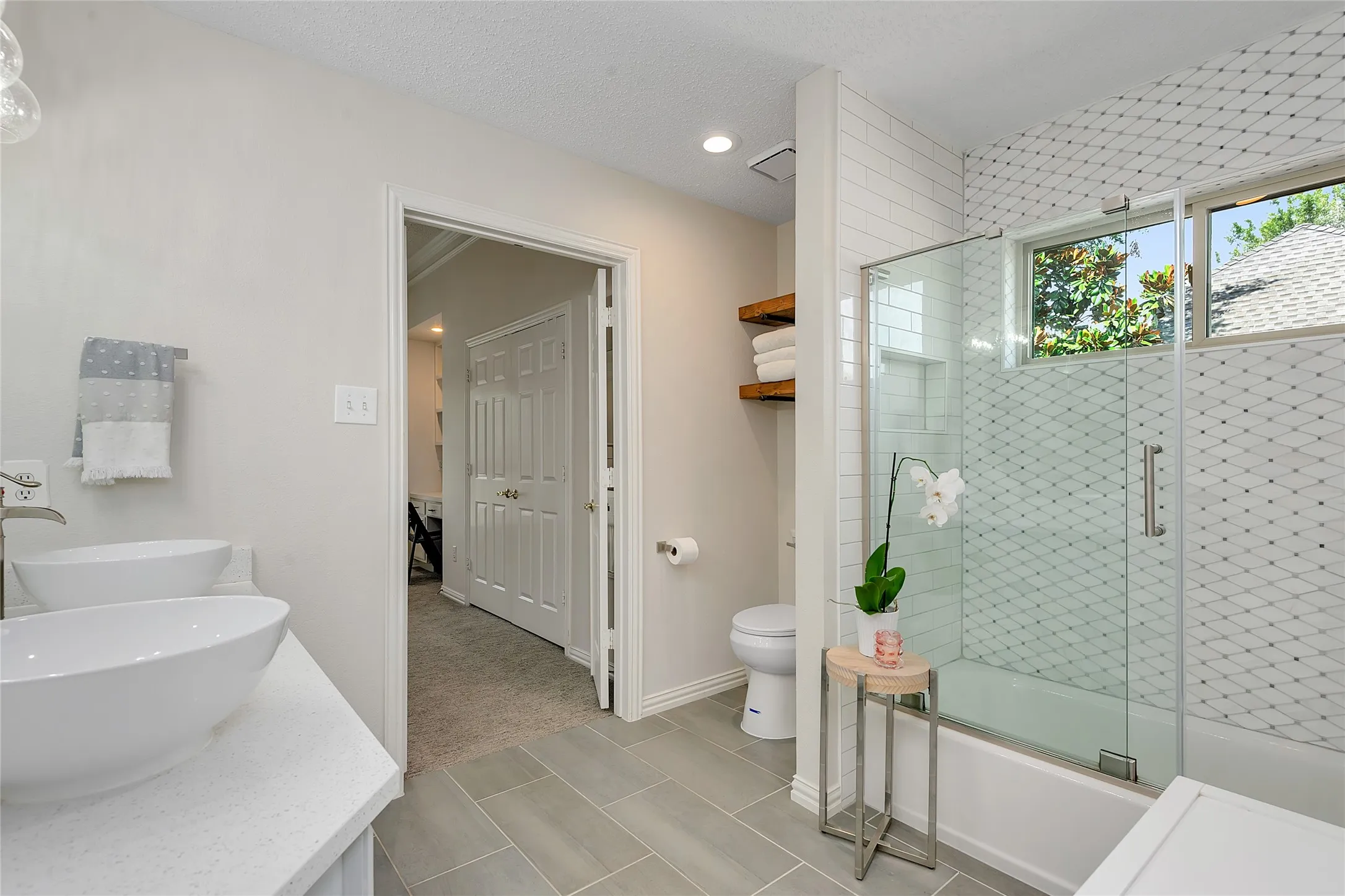 Bathroom featuring a dual vessel sink vanity with light-toned countertops, a glass-enclosed shower-over-tub with patterned tile, wood-finish floating shelving, and light-toned tile flooring