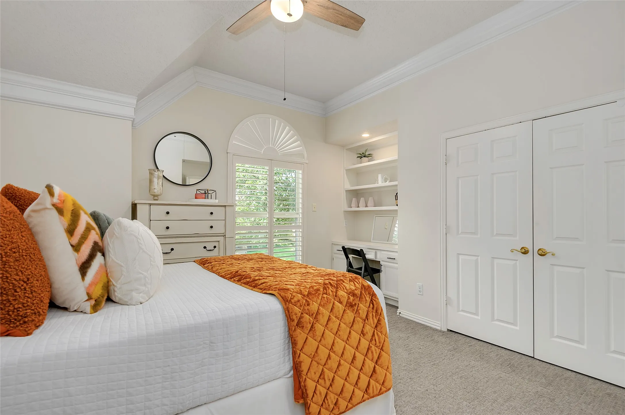 Vaulted ceiling bedroom featuring crown molding, neutral carpeting, a built-in desk with shelving, and plantation shutters on the arched window