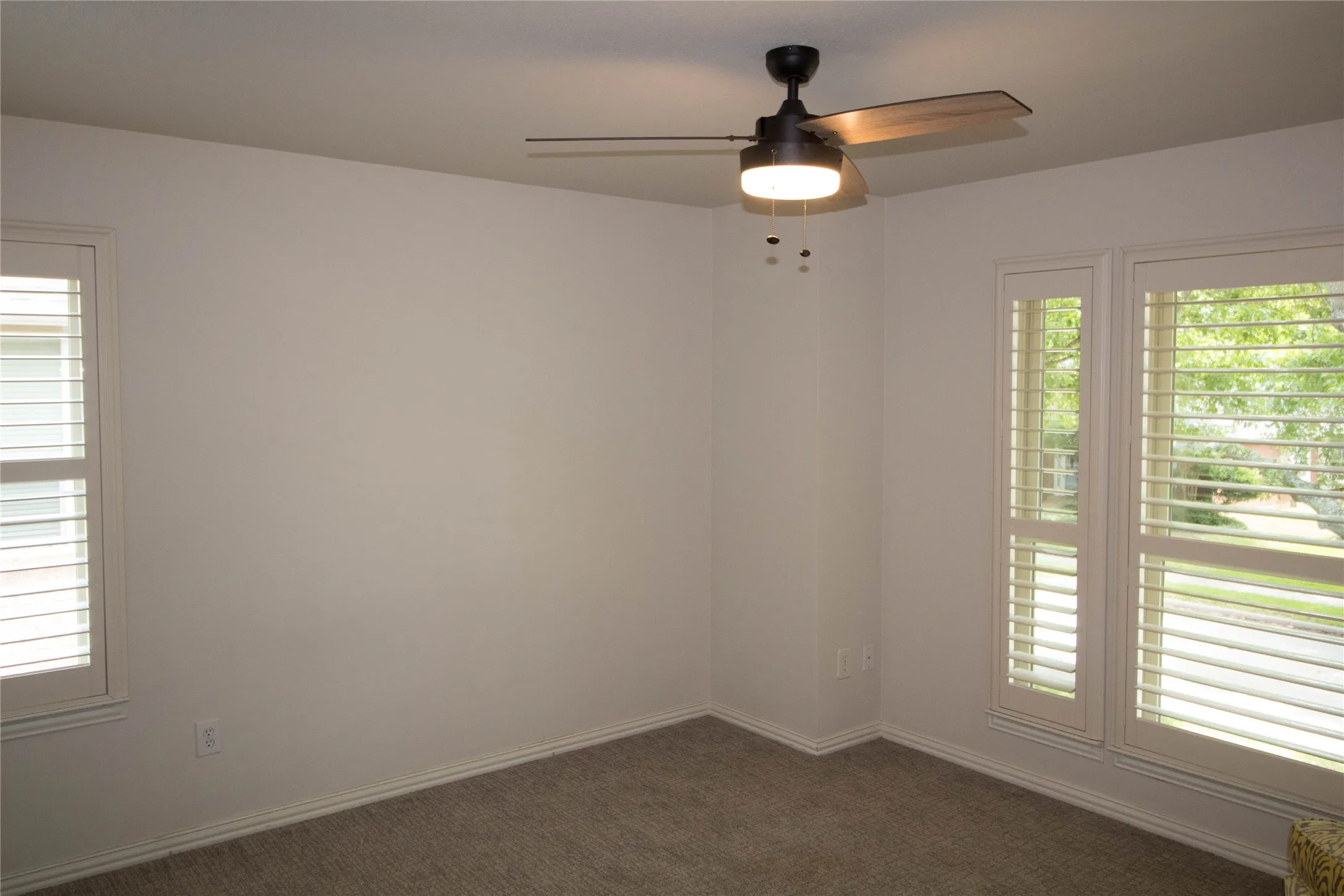 Bright room featuring two large windows with plantation shutters, a ceiling fan with light fixture, and neutral carpeting