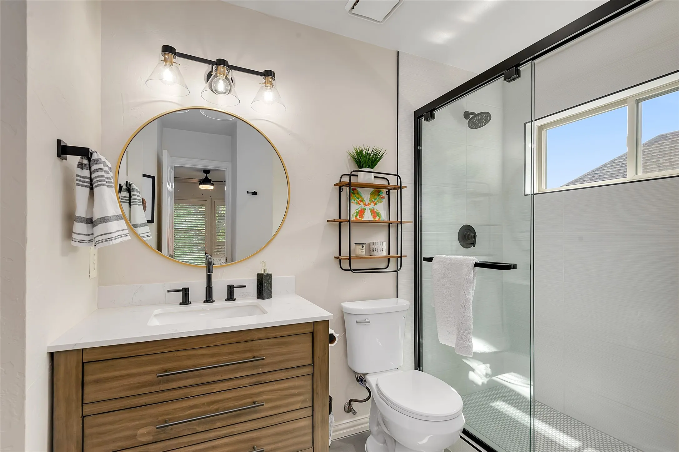 Bathroom featuring a glass-enclosed shower with black trim, a wood-finish vanity with a white countertop, and a circular gold-framed mirror