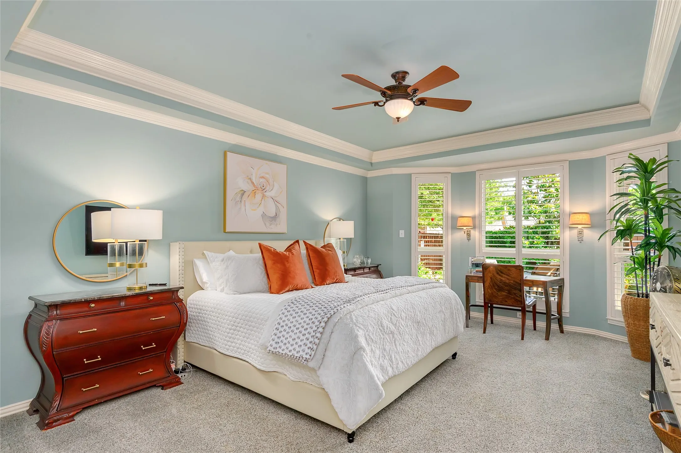 Recessed tray ceiling with crown molding, ceiling fan with integrated lighting, bay window with plantation shutters, wall-mounted sconces, and light-toned carpeting