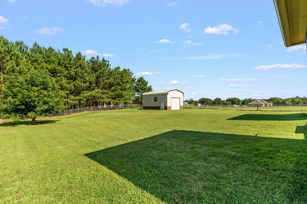 Expansive green lawn featuring a detached metal garage building with a roll-up door, a chain-link fence, mature trees along the perimeter, and clear skies