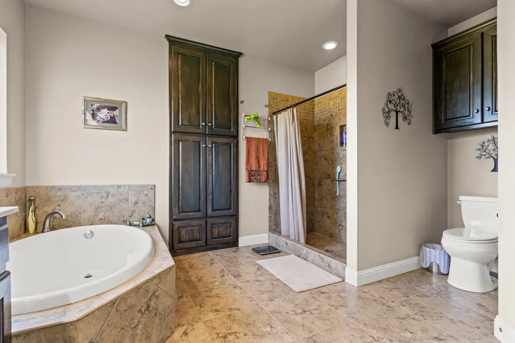 Spacious bathroom featuring a built-in soaking tub with stone tile surround, a glass-enclosed shower with stone tile, dark wood-finish cabinetry, and neutral-toned floor tile