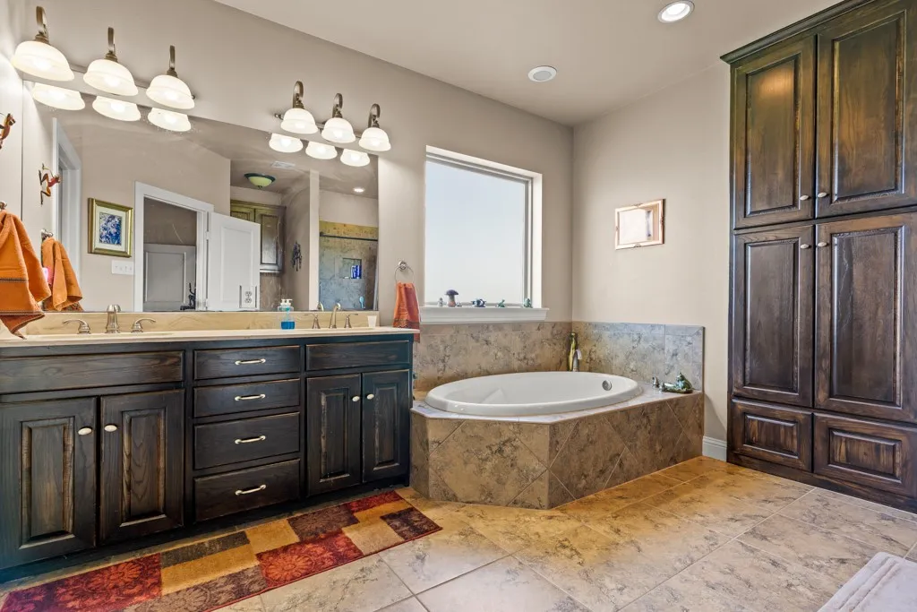 Spacious bathroom featuring a built-in soaking tub with tiled surround, dual vanity with dark wood cabinetry and light countertop, large wall mirror with dual light fixtures, and a tall built-in storage cabinet