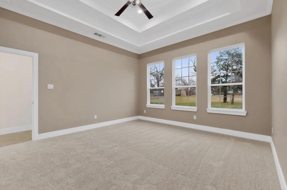 Spacious room featuring three large windows, a tray ceiling with a ceiling fan, plush carpeting, and light neutral wall tones
