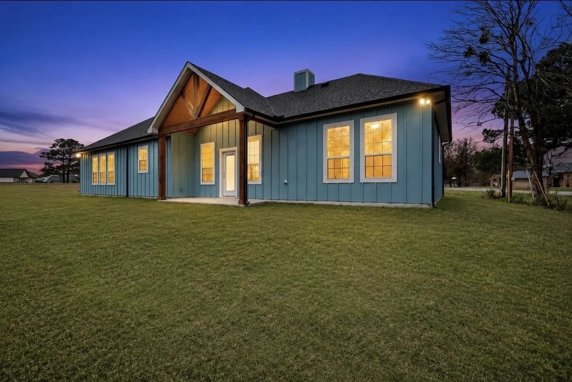 Rear exterior featuring blue vertical board and batten siding, a covered patio with wood beams, a dark shingle roof, and multiple windows