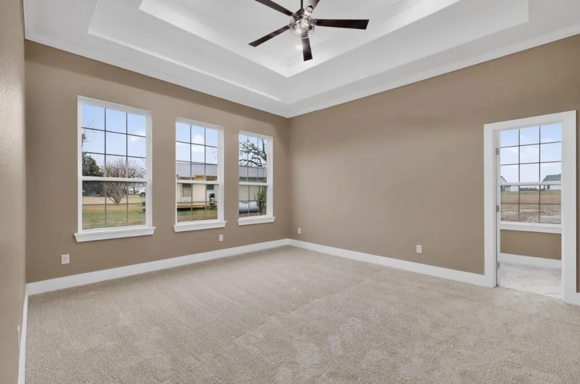 Spacious room featuring a tray ceiling with a ceiling fan, three large grid-pattern windows, neutral-toned walls, plush carpeting, and crisp white trim