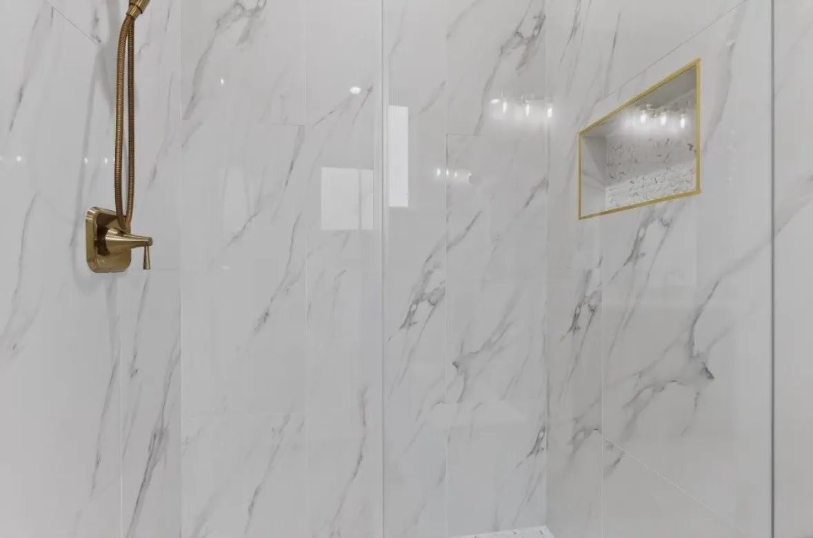 Shower enclosure featuring large format white tile with grey veining, a built-in niche with gold-toned trim, a gold-toned shower fixture, and clear glass panels
