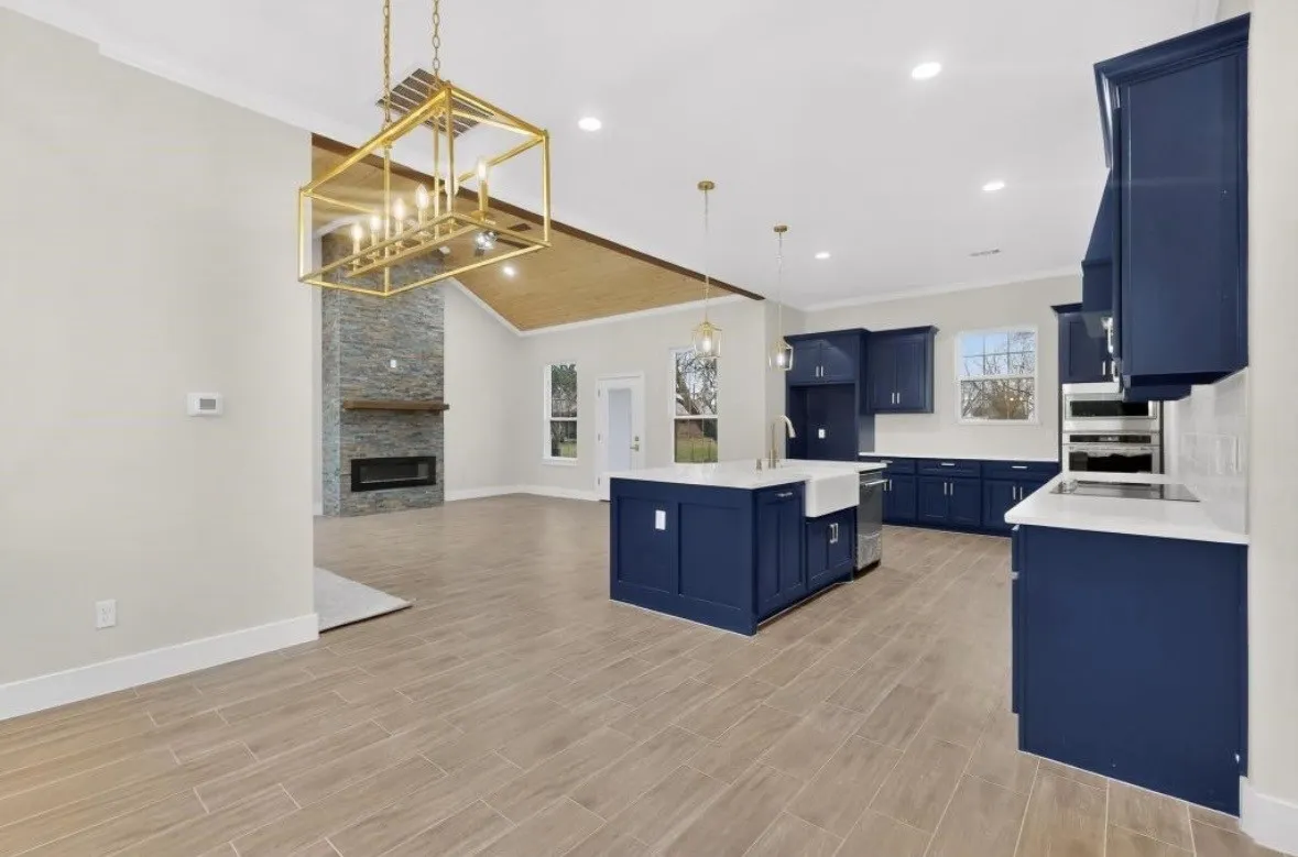 Open-concept interior featuring light wood-finish flooring, a contemporary kitchen with navy cabinetry and white countertops, a central island with a farmhouse sink, and a stone-clad fireplace with a wood mantle