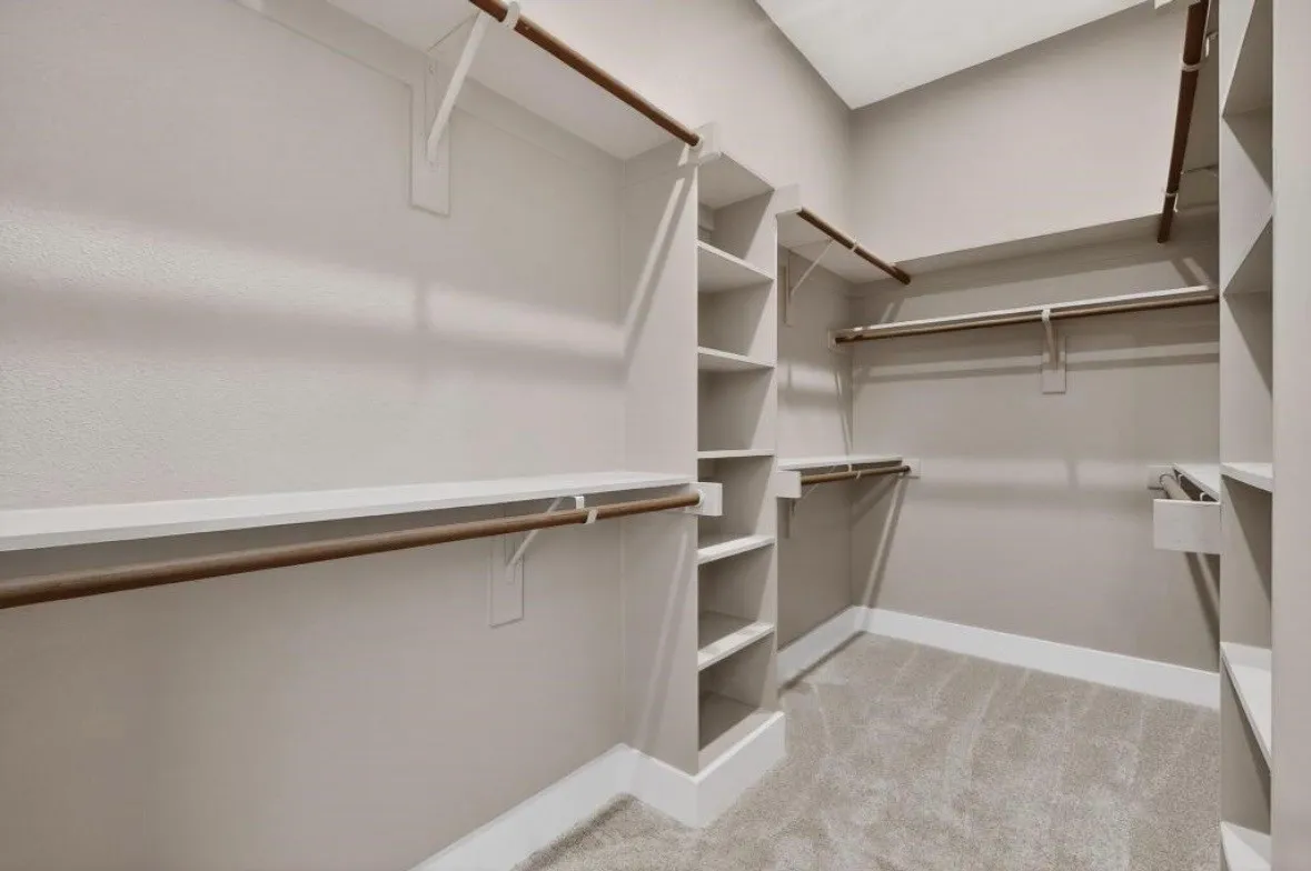 Walk-in closet featuring light gray walls, carpet flooring, multiple wooden hanging rods, white shelving units, and white baseboards