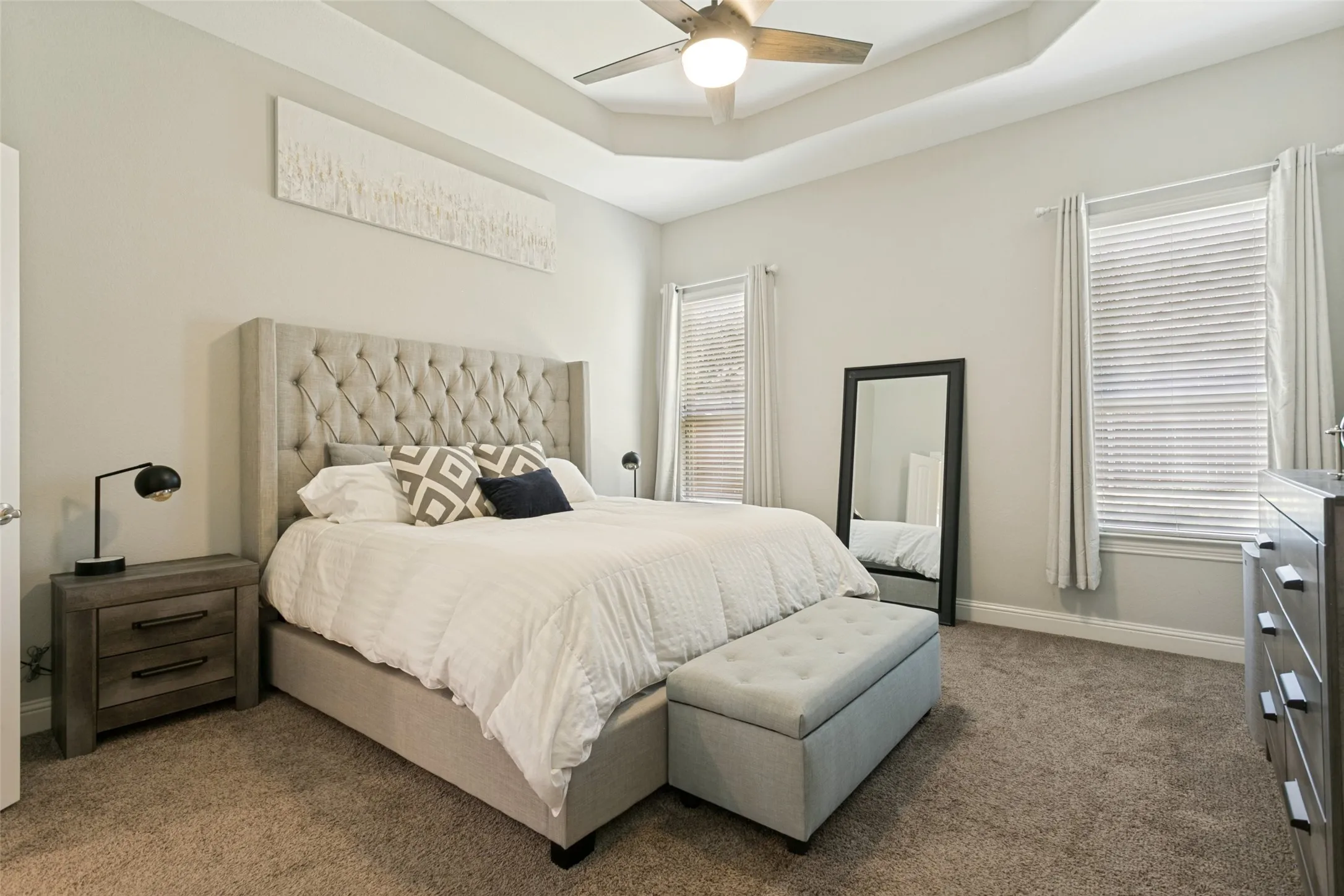 Tray ceiling with ceiling fan, two windows with blinds and curtains, and neutral carpet flooring