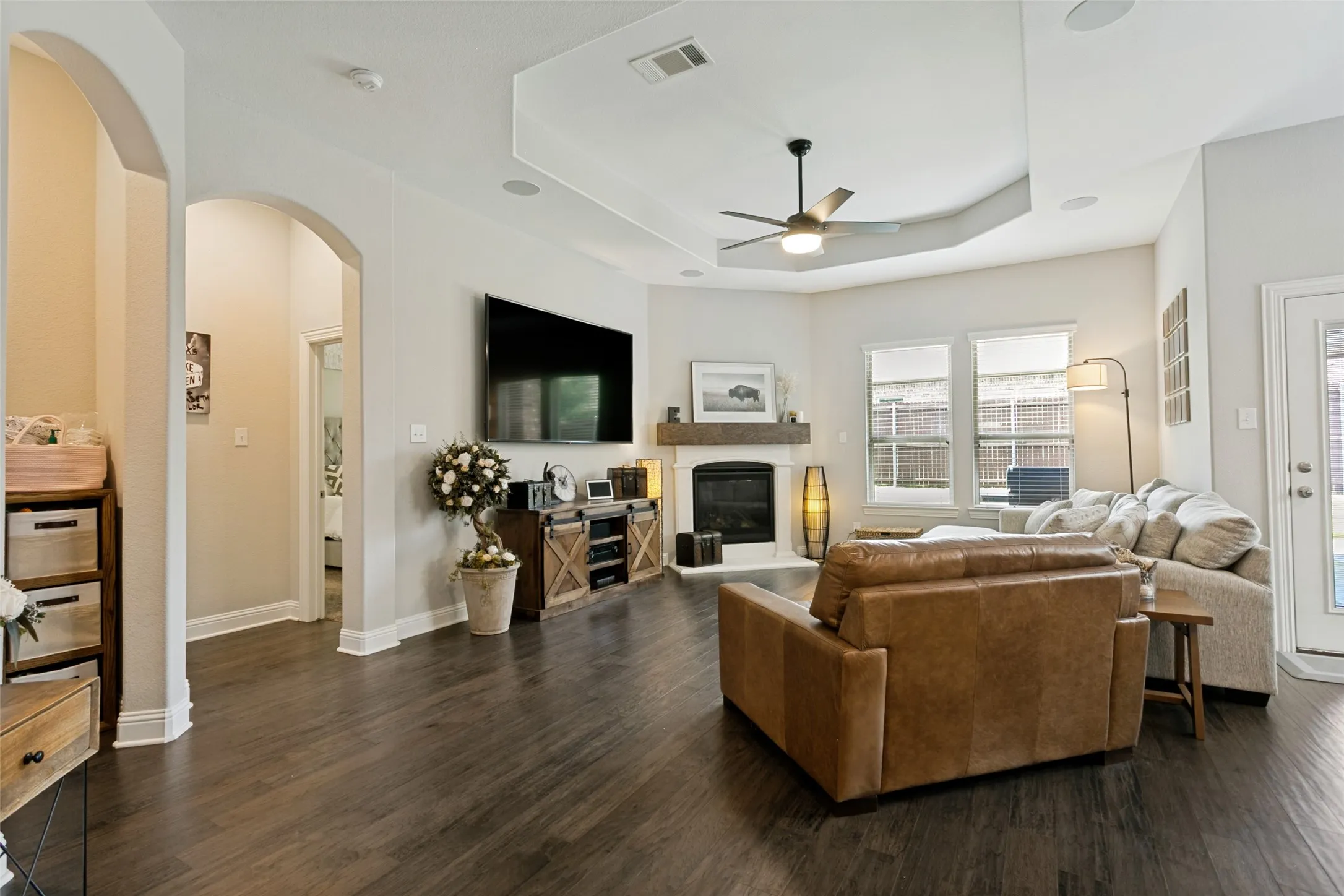 Living room featuring dark wood-finish flooring, a tray ceiling with a ceiling fan, a white fireplace with a mantel, and three windows providing natural light