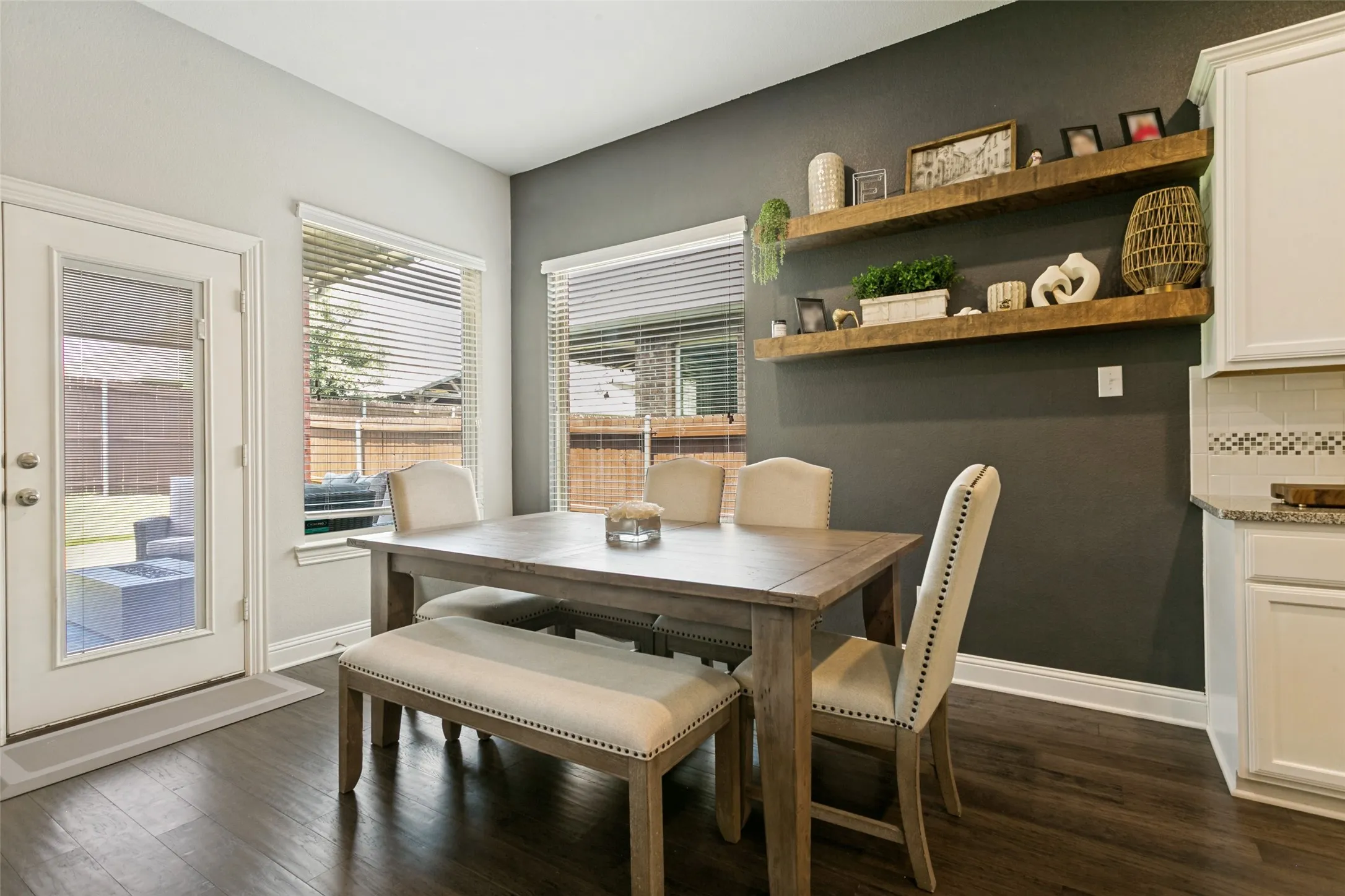 Dining area featuring rich wood-finish flooring, a full-lite exterior door, dual windows with blinds, and a contrasting accent wall with two wood floating shelves