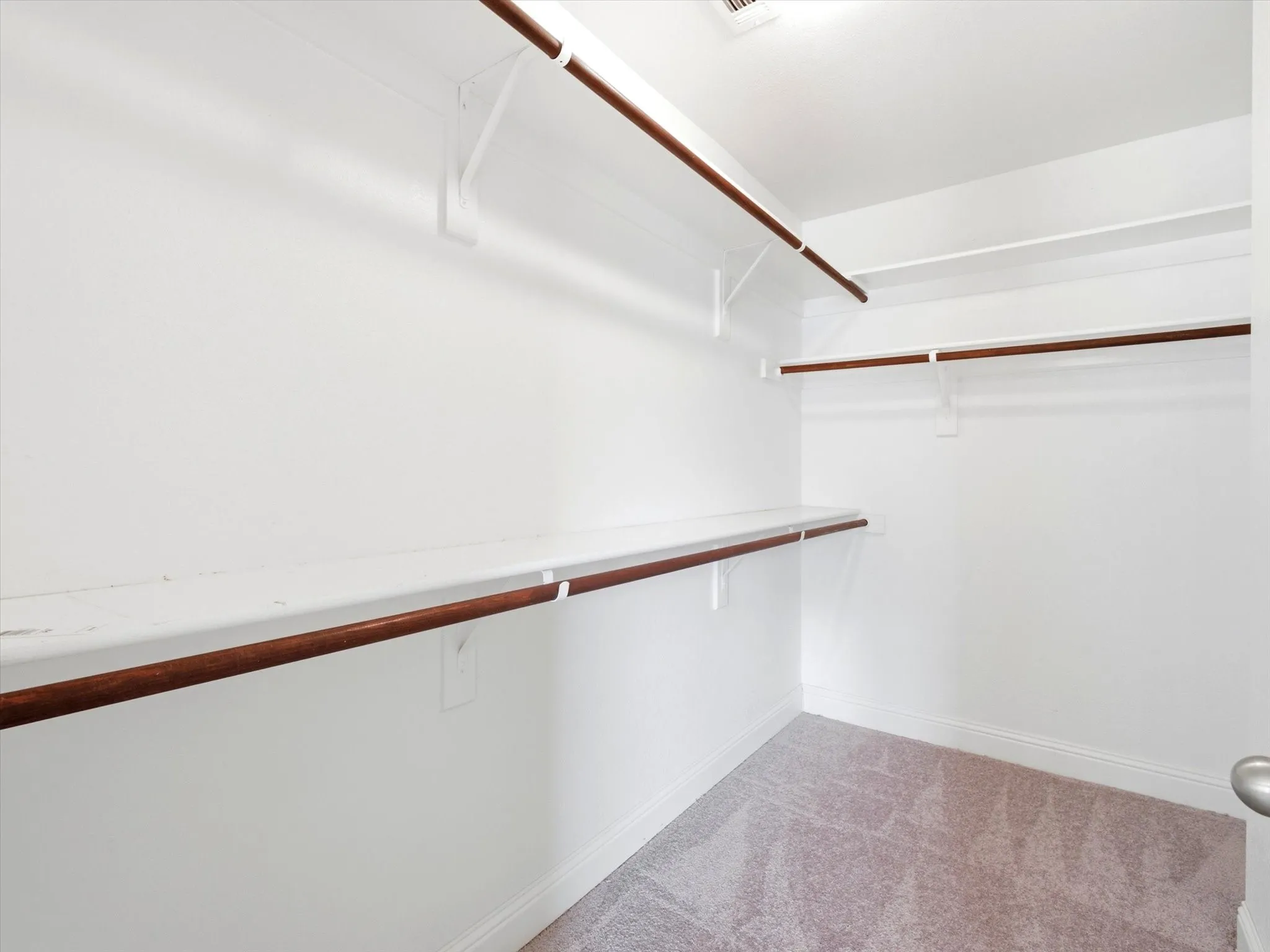 Spacious walk-in closet featuring multiple wooden rod systems at varying heights, white painted walls, and neutral carpeting for optimal storage and organization