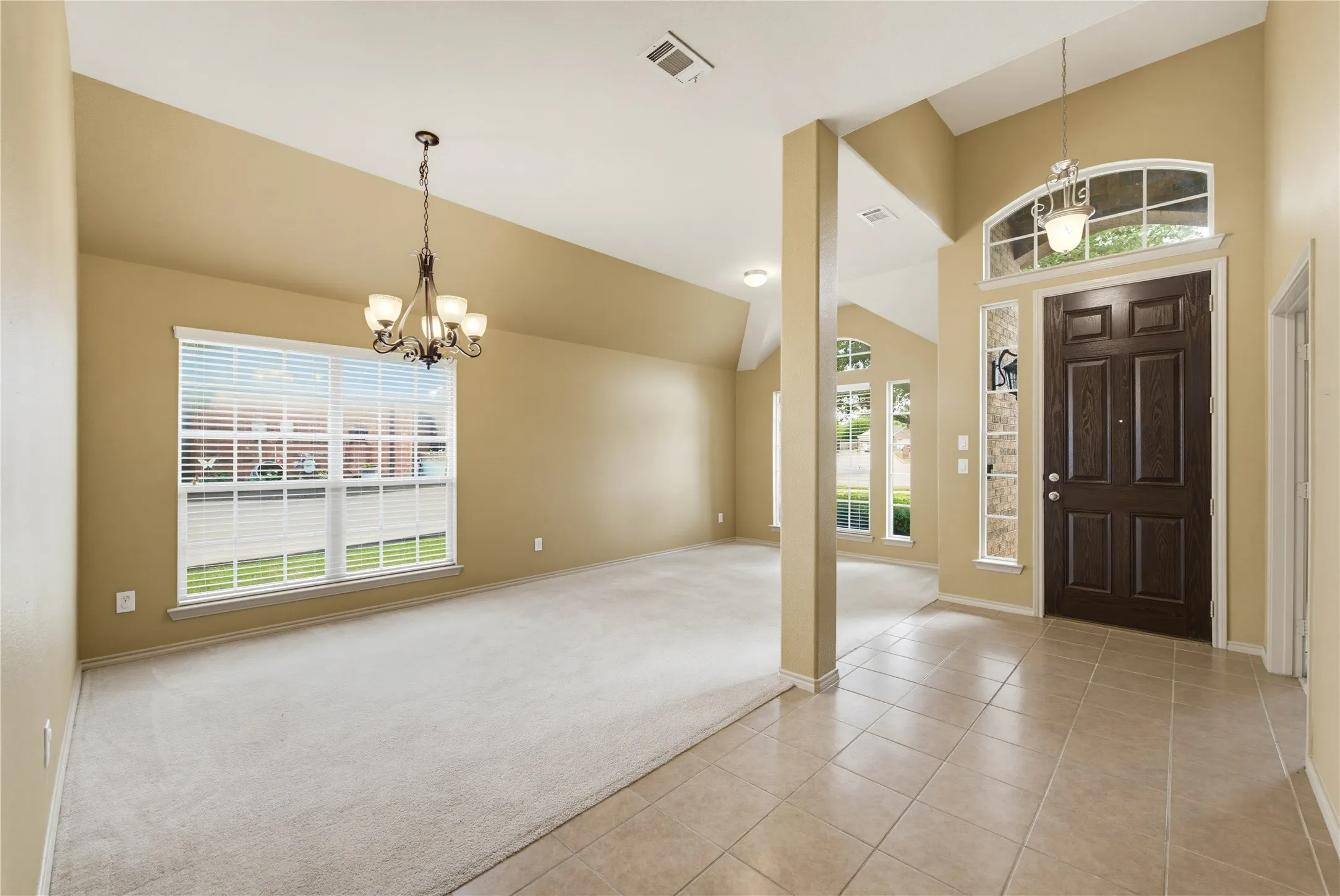 Entryway and adjoining formal dining area at 11 Beechcreek Drive in Edgecliff Village, TX 76134, near Fort Worth. The unfurnished interior features high ceilings, neutral beige walls, a tiled foyer, light carpeting, decorative columns, and hanging light fixtures. A dark wood front door is framed by a narrow sidelight and an arched transom window, while large windows with white blinds brighten the dining area.