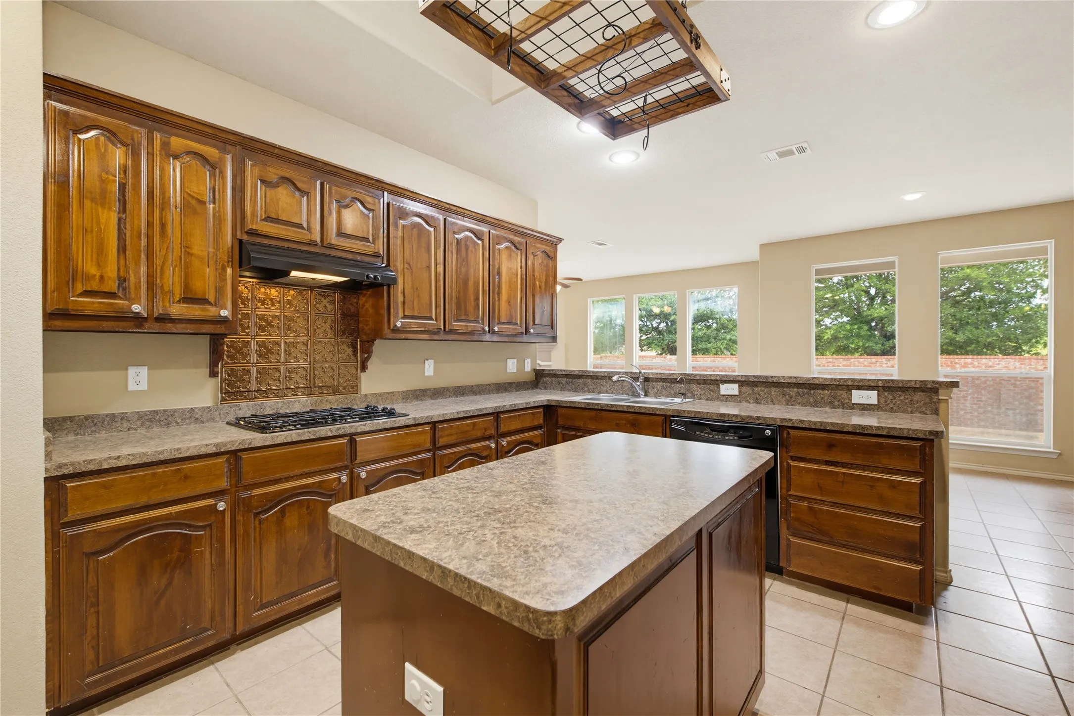 Kitchen at 11 Beechcreek Drive in Edgecliff Village, TX 76134, near Fort Worth. The space features stained wood cabinetry, a central island, patterned countertops, a gas cooktop with a decorative metallic backsplash, a double sink, and a black dishwasher. Tile flooring, recessed lighting, and an overhead pot rack complete the kitchen, which overlooks an adjoining area with large windows facing the backyard.