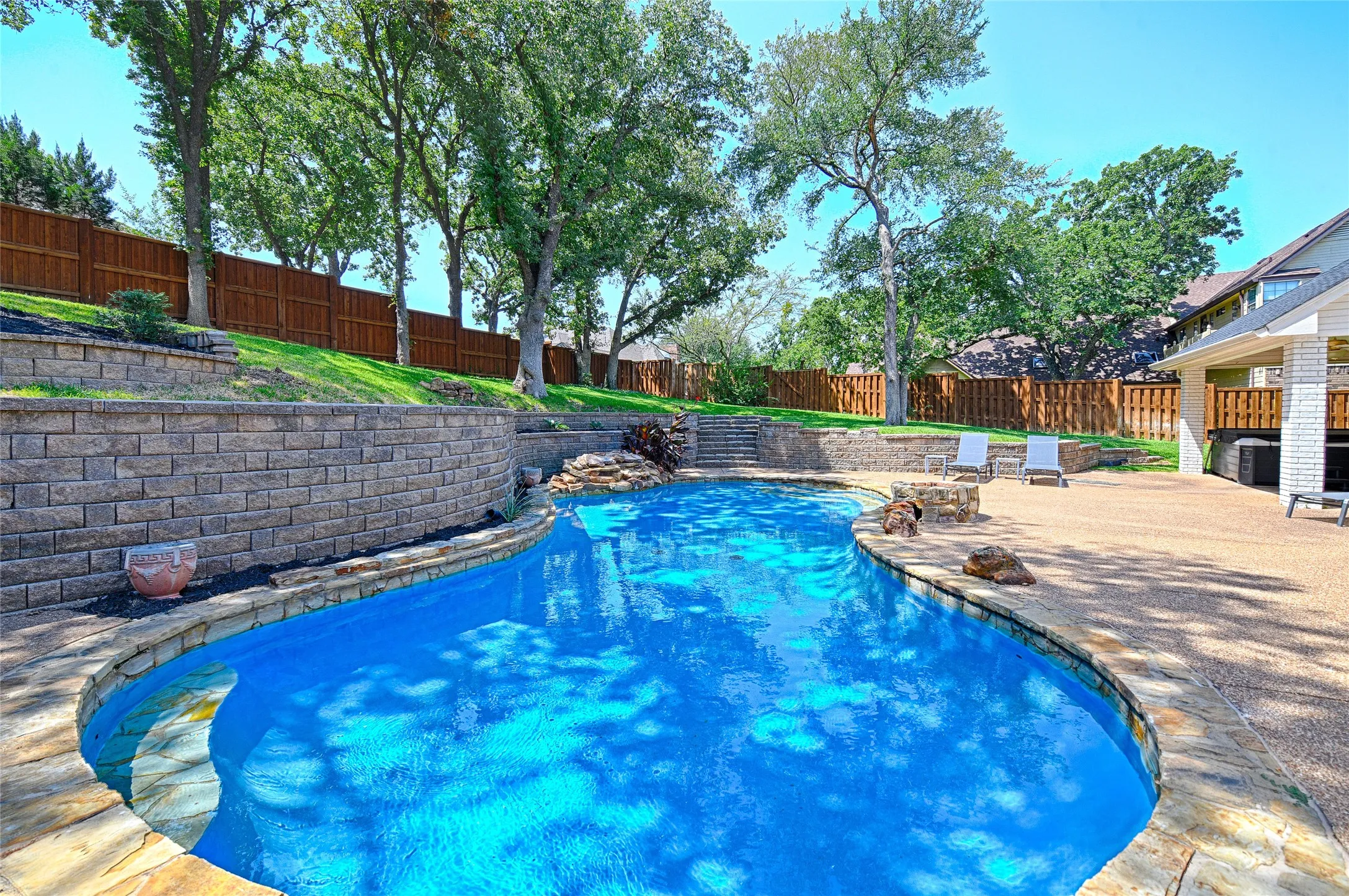 Curved swimming pool with stone coping, a tiered retaining wall, and a waterfall feature