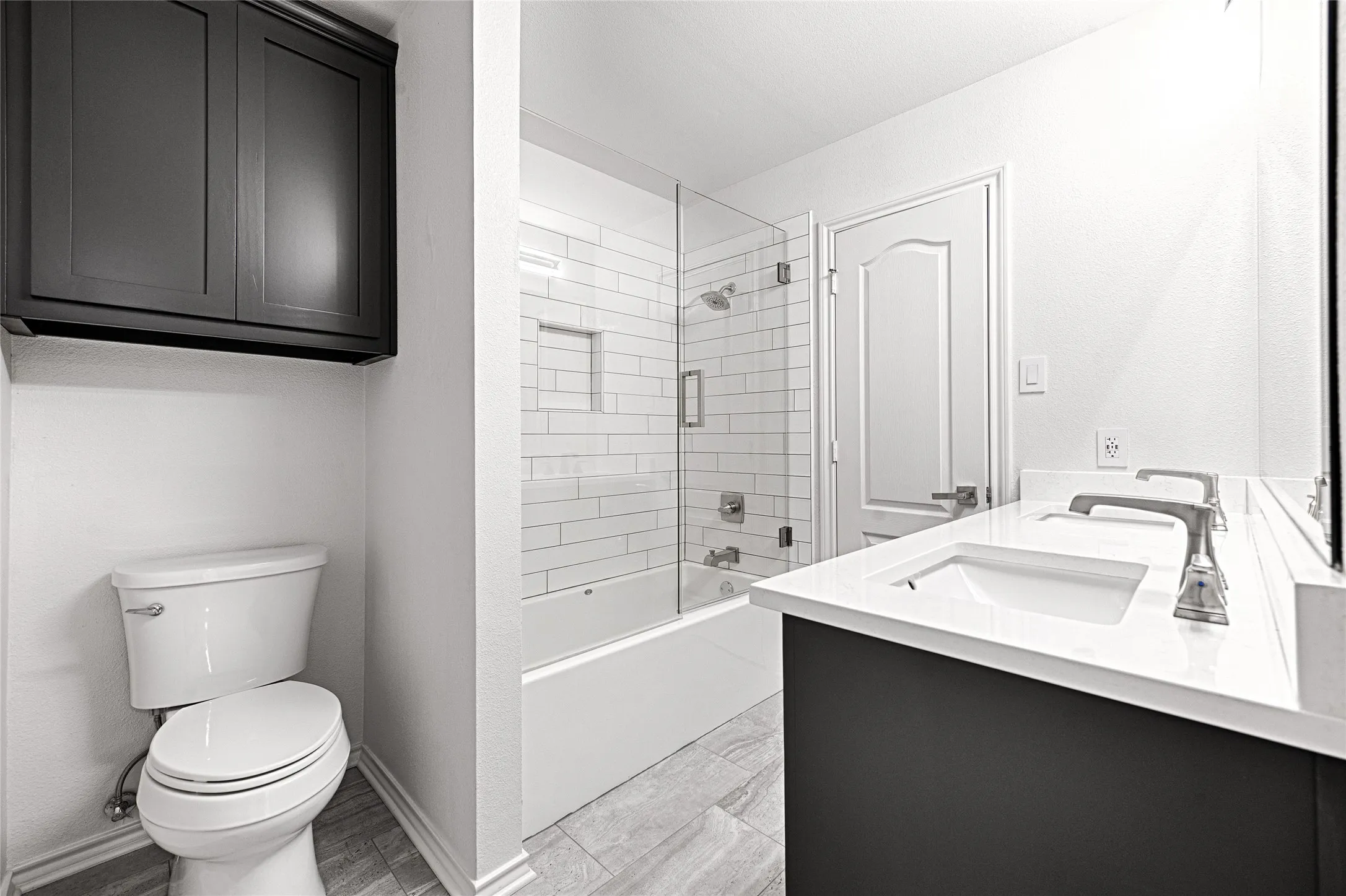 Bathroom featuring a dual vanity with undermount sinks and light-toned countertops, a glass-enclosed shower over a tub with subway tile surround, a built-in shower niche, and a wall-mounted dark cabinetry above the commode