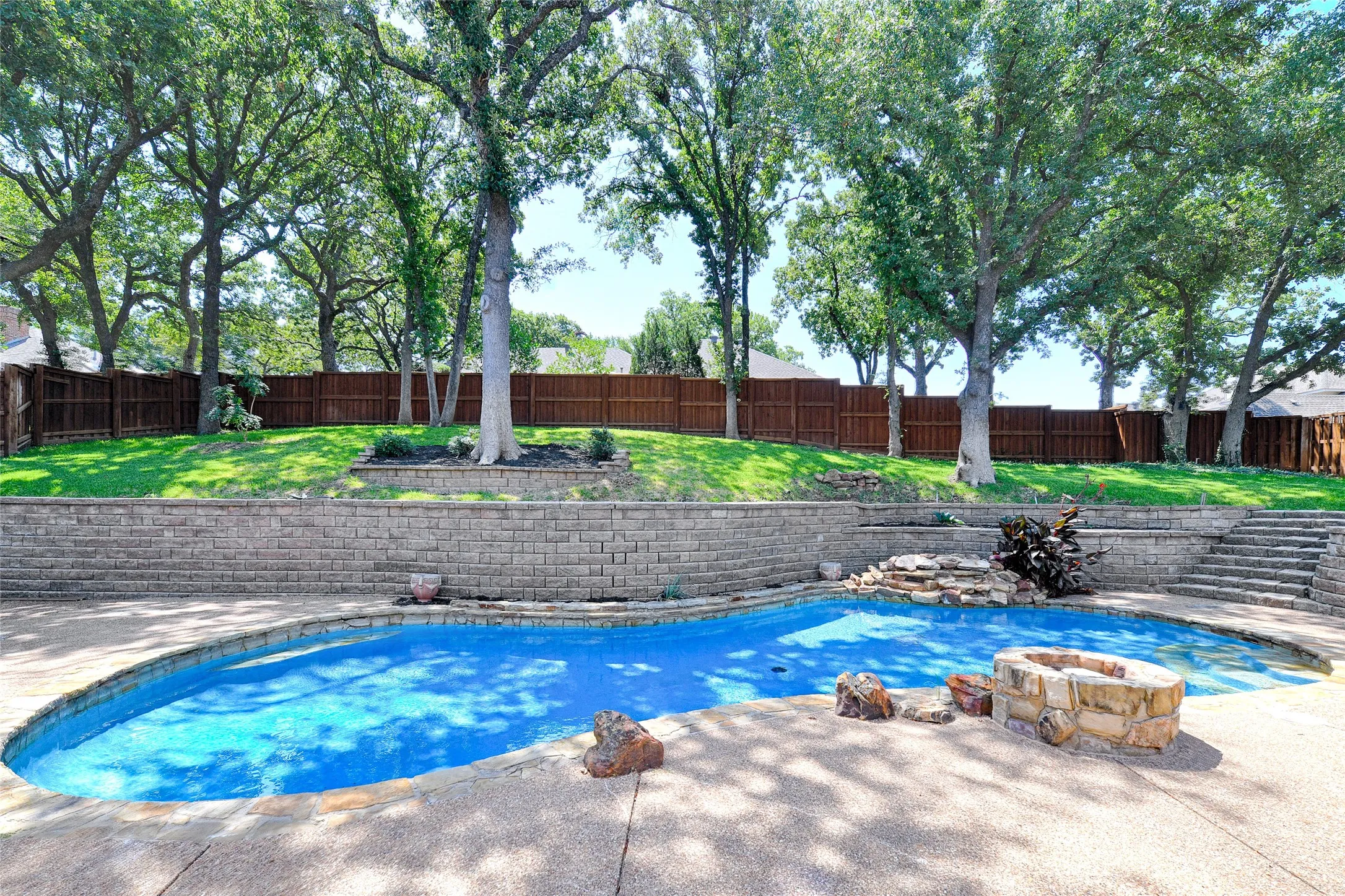 Expansive backyard featuring a freeform swimming pool with a rock waterfall feature, a stone fire pit, extensive concrete decking, and tiered landscaping with a retaining wall