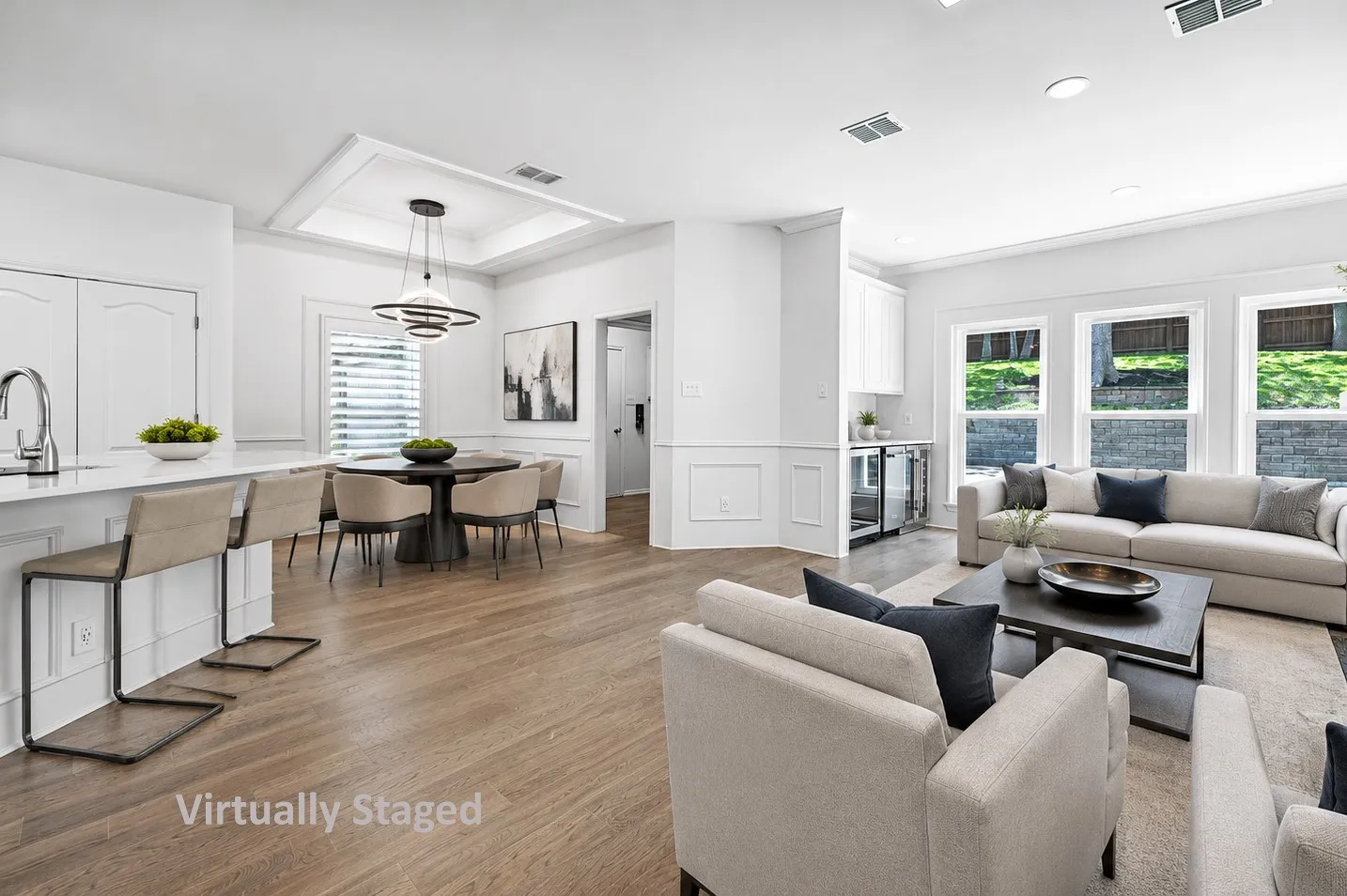 Virtually Staged: Open-concept living area with wood-finish flooring, a kitchen island with seating, and a dining area featuring a tray ceiling and contemporary pendant lighting