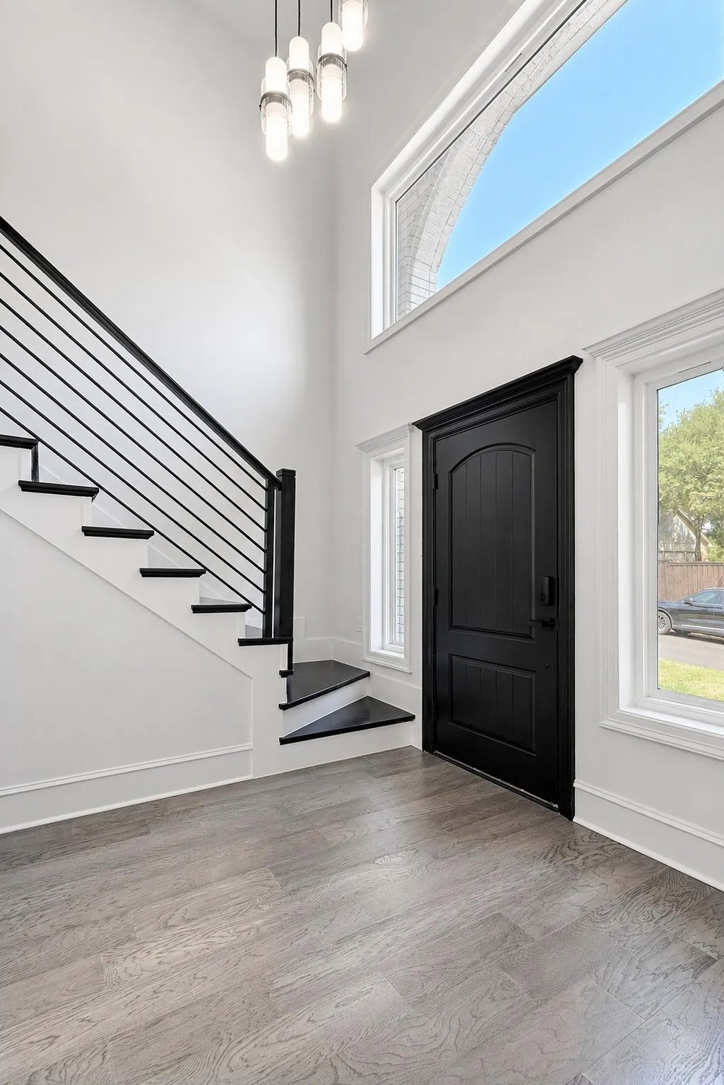 Two-story entryway featuring a contemporary chandelier, high ceilings, wood-finish flooring, a black paneled door, and a staircase with a horizontal metal railing