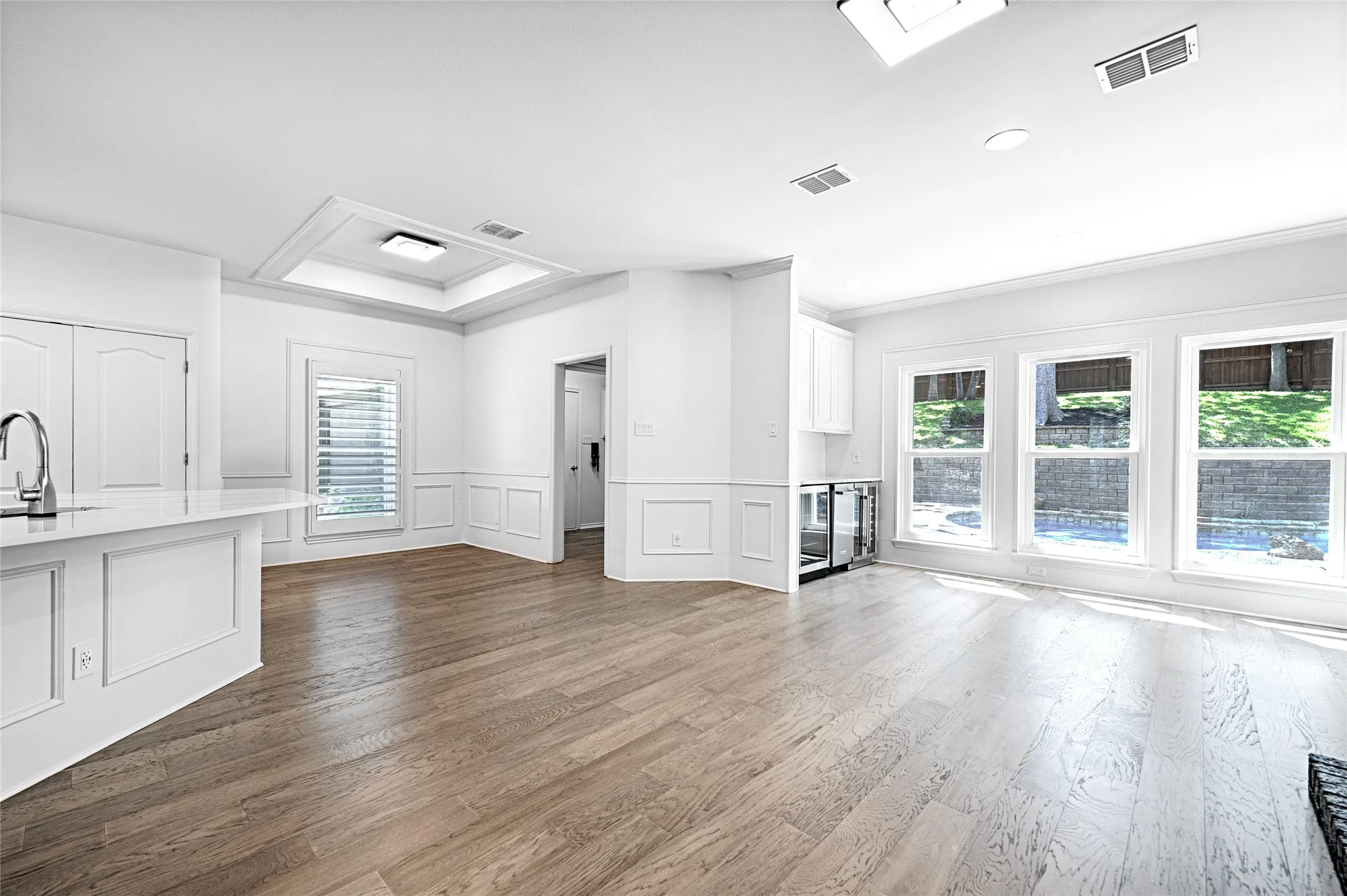 Open-concept living area with wood-finish flooring, white wainscoting, and large windows overlooking a terraced exterior with pool & patio access