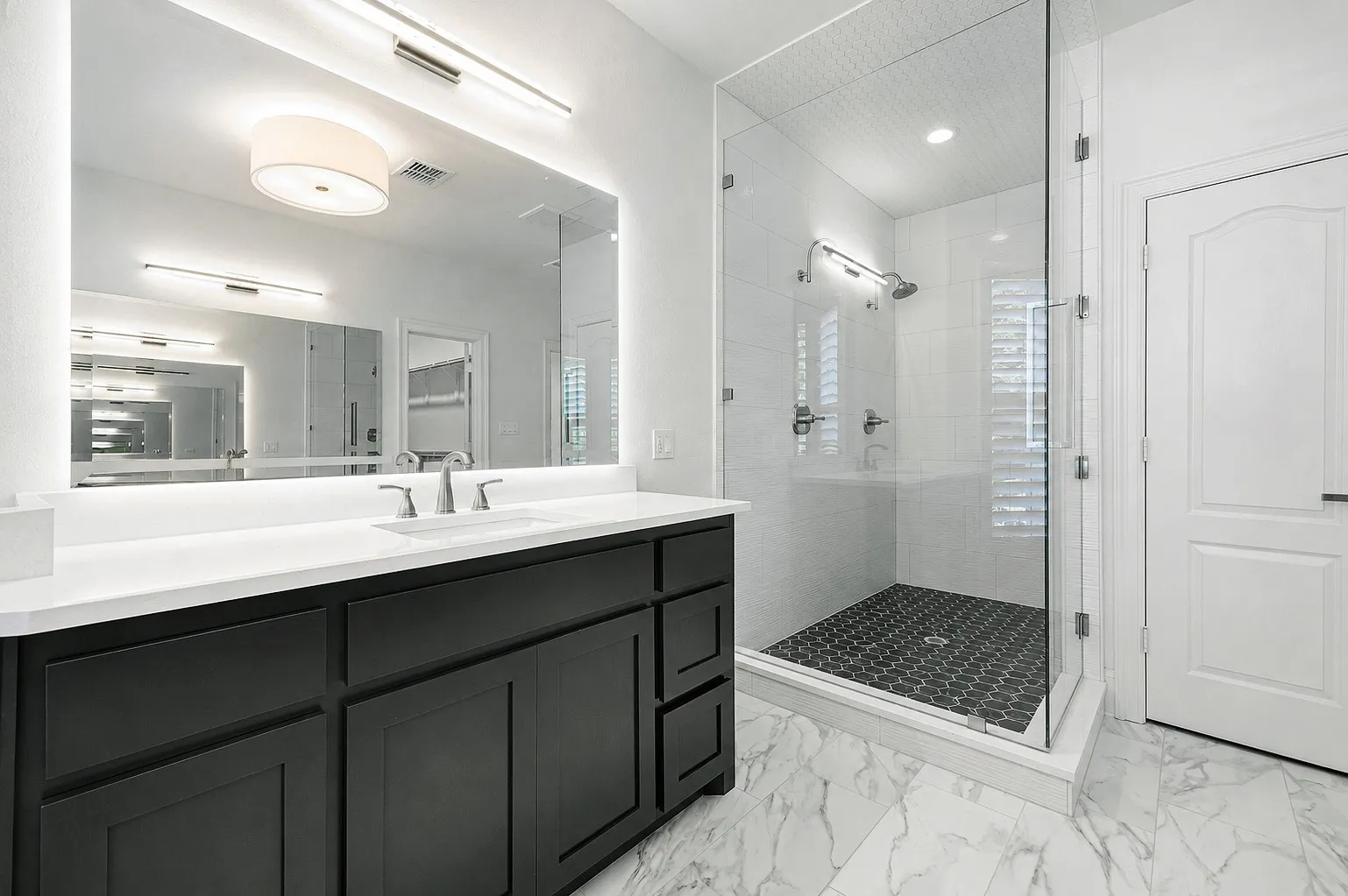 Bathroom featuring a frameless glass shower enclosure with hex tile flooring, a dark wood-finish vanity with a white countertop, an integrated rectangular sink, and a large illuminated mirror