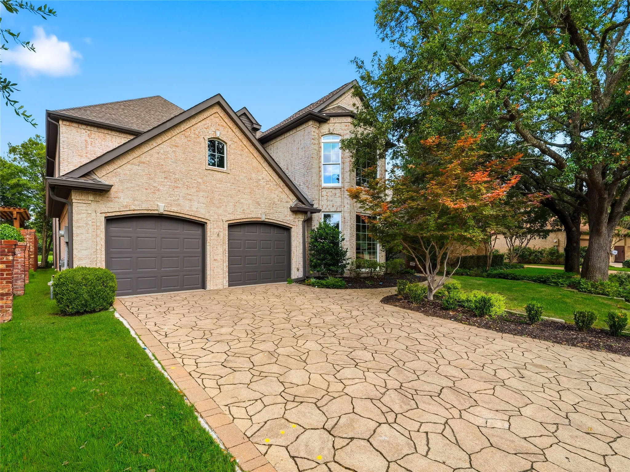 Brick exterior featuring a two-car garage with arched doors, a patterned stone driveway, and mature landscaping