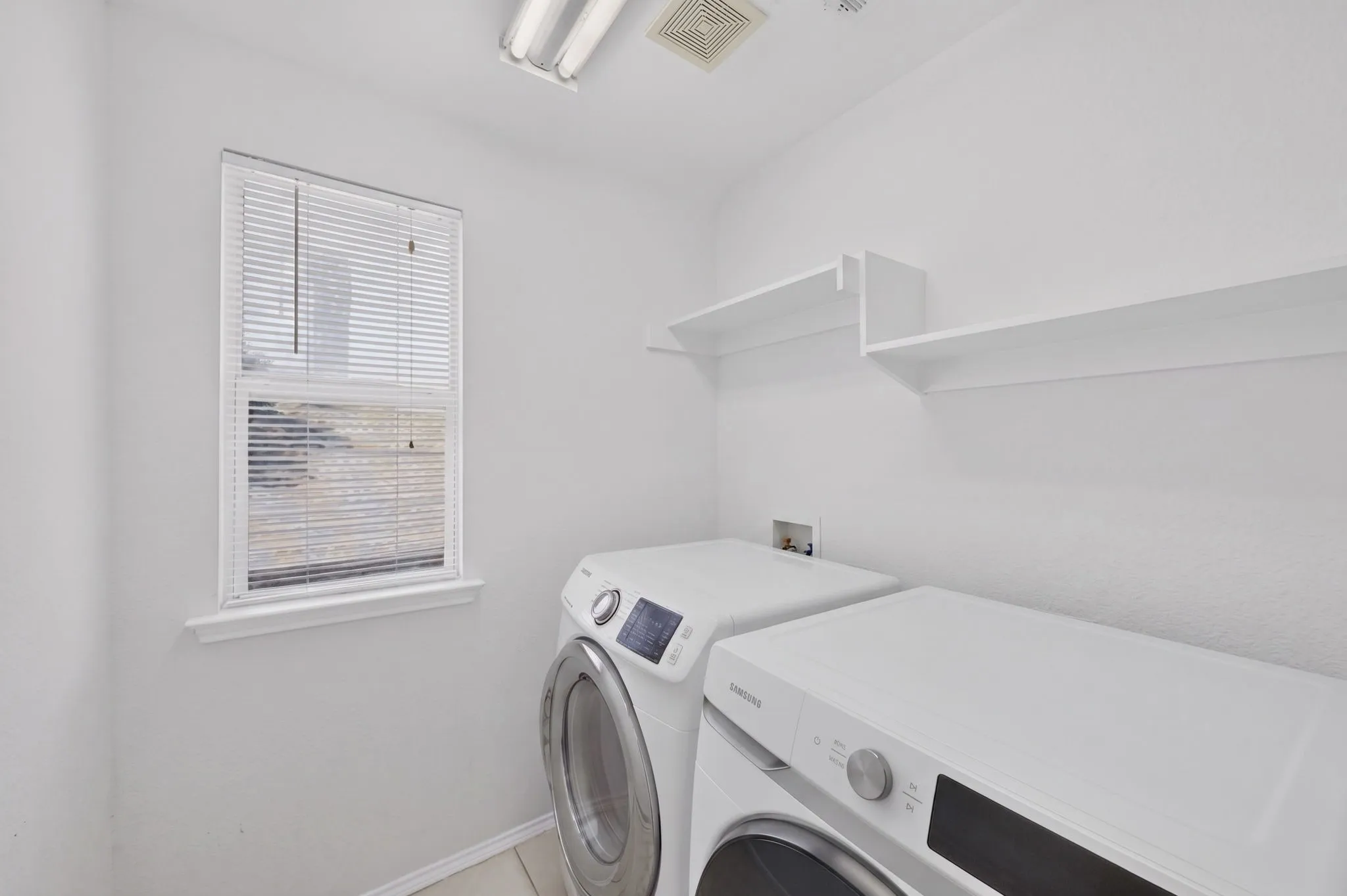 Dedicated laundry area featuring white walls, light-colored tile flooring, a window with horizontal blinds, and built-in shelving