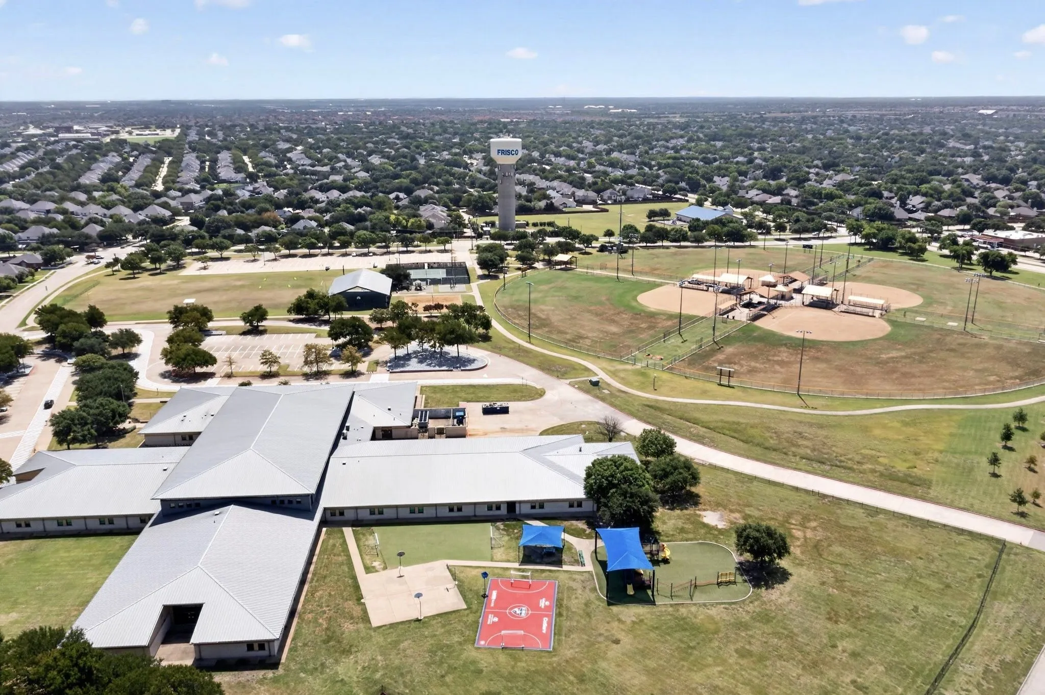 Expansive overhead view showcasing a large community building with a metal roof, multiple sports fields, and a water tower