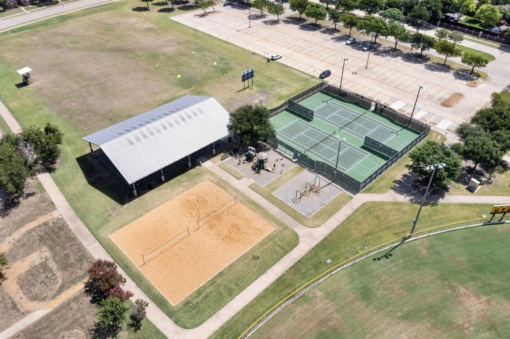 Community recreational facility featuring a large covered pavilion, multiple tennis courts with fencing and lighting, a sand volleyball court, and a nearby open field