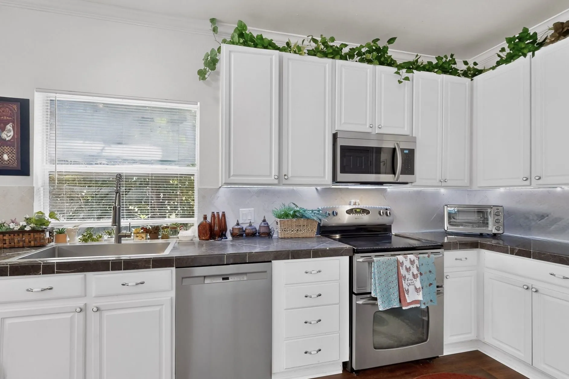 Well-appointed kitchen featuring white cabinetry, stainless steel appliances, a double basin sink, tiled countertops, and under-cabinet lighting