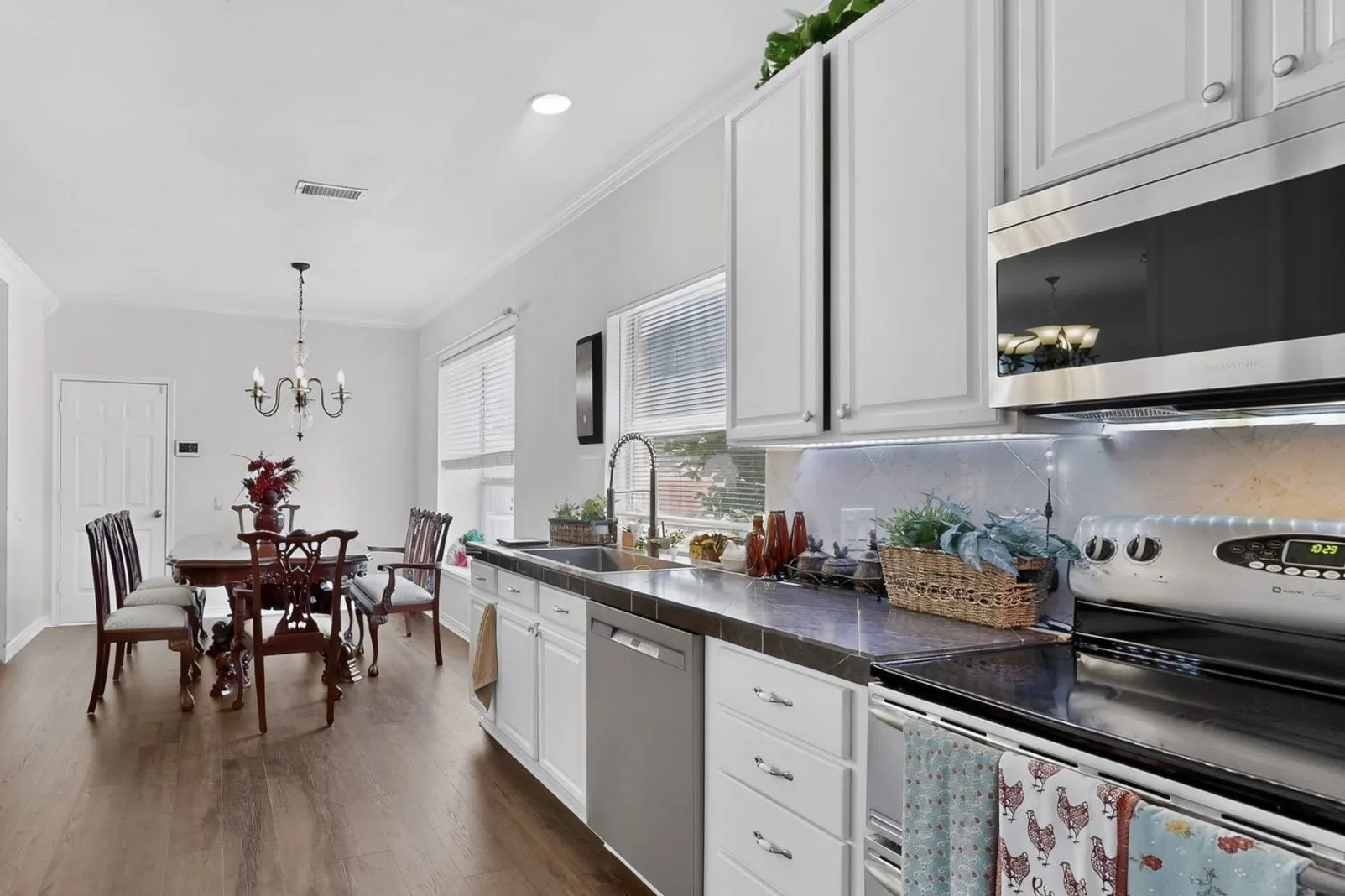 Bright kitchen and dining area featuring wood-finish flooring, white cabinetry with silver hardware, an undermount sink, stainless steel appliances, and crown molding